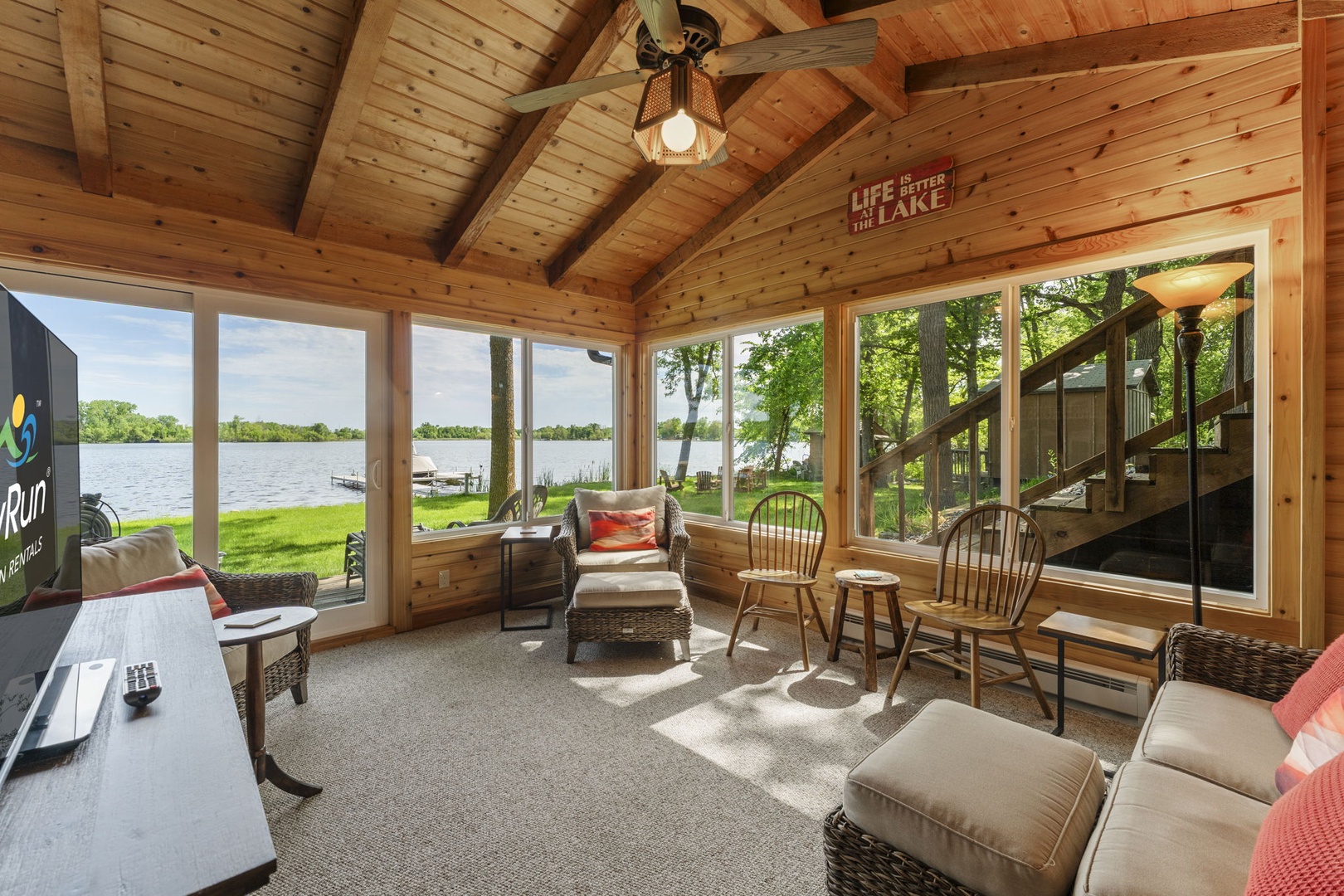 A bright lakeside sunroom features vaulted wood ceilings, panoramic windows framing the sparkling lake, and comfortable seating perfect for unwinding after a day on the water.