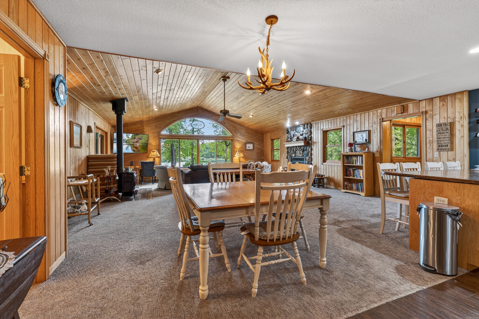 Gather around the rustic dining table in this cozy, wood-paneled retreat. Natural light pours through large windows, while a charming antler chandelier sets a warm, inviting tone overhead.