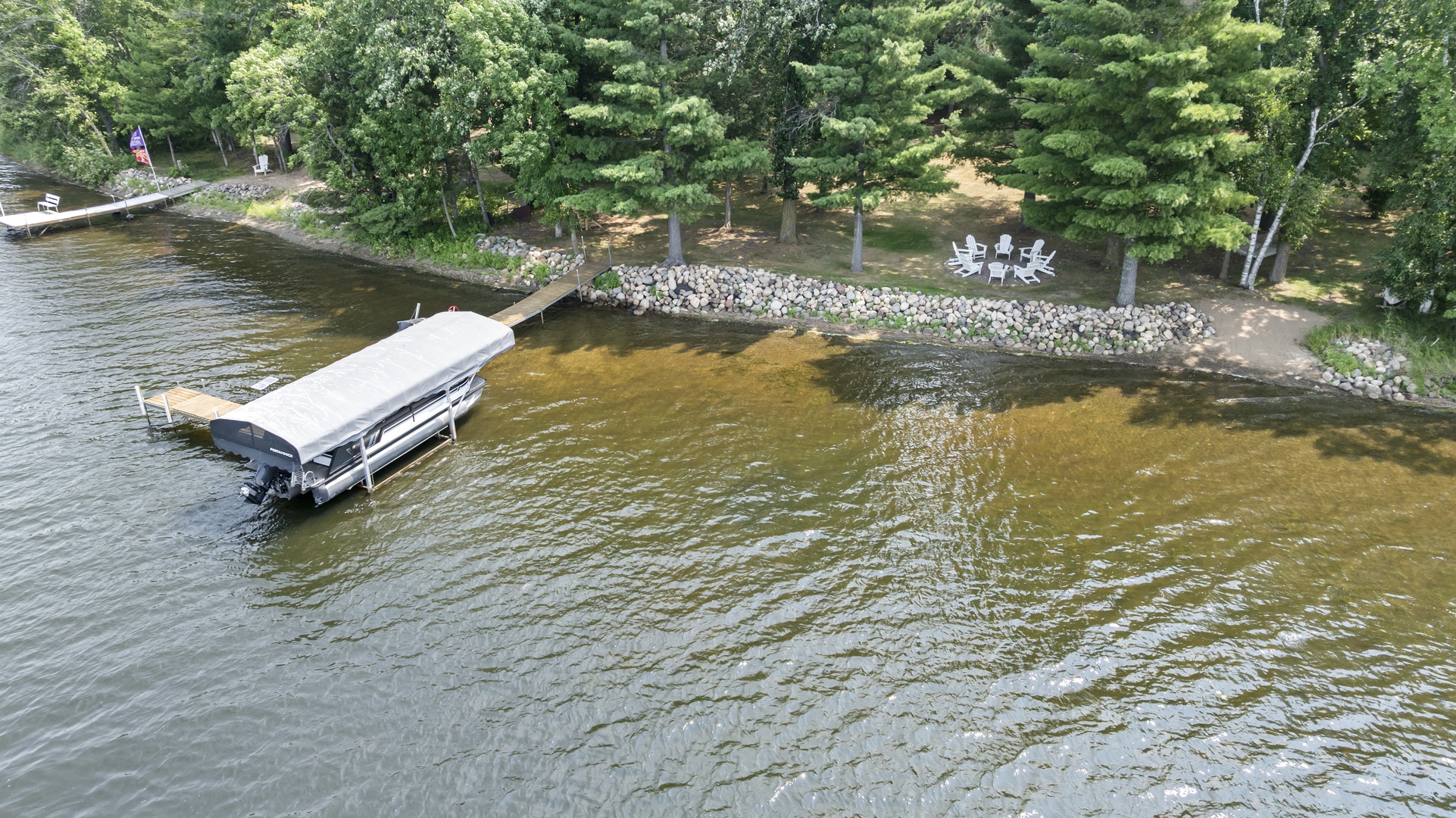 Aerial view of lakefront property featuring a private dock with boat lift and waterfront seating area bordered by natural stone, surrounded by lush wooded landscape.
