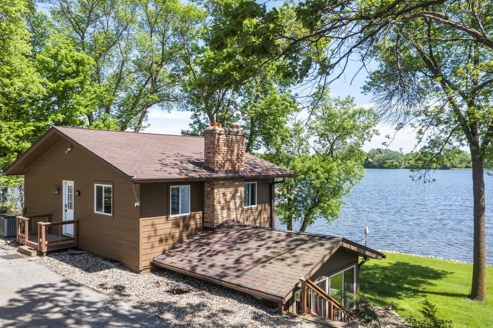 Lakefront cabin nestled among mature trees with a charming brick chimney, wooden deck, and private waterfront access on a tranquil summer day.