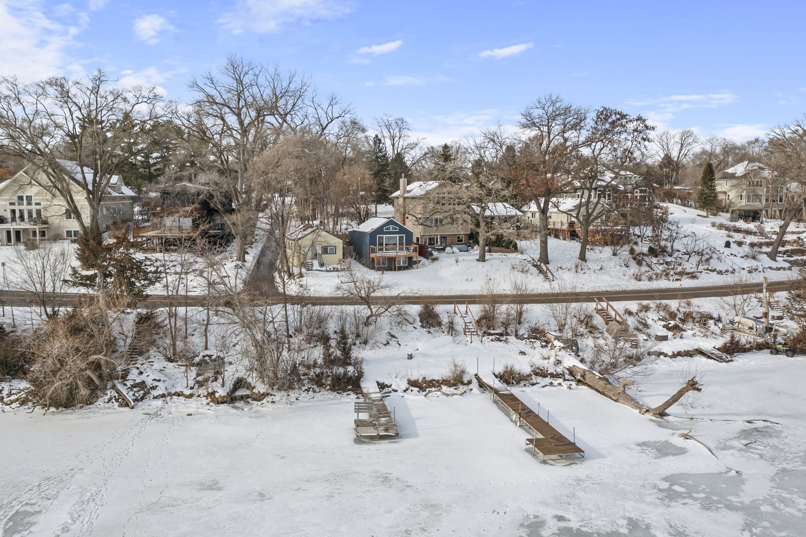 A peaceful lakefront neighborhood blanketed in pristine snow, featuring charming homes nestled among bare winter trees beneath a bright blue sky.
