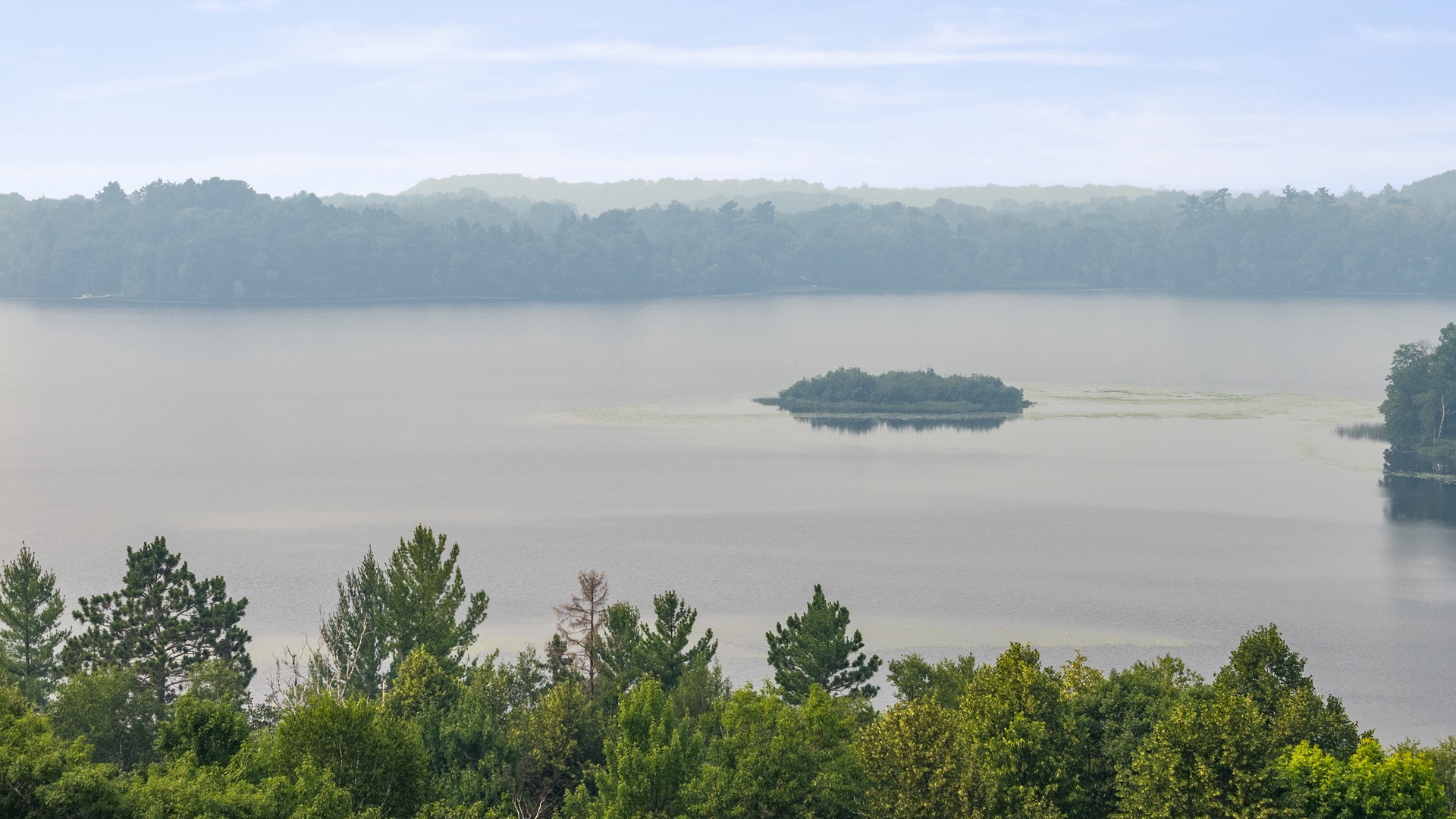 A serene lake with forested islands and gentle morning mist drifting over calm waters, surrounded by lush green hills and abundant natural beauty.