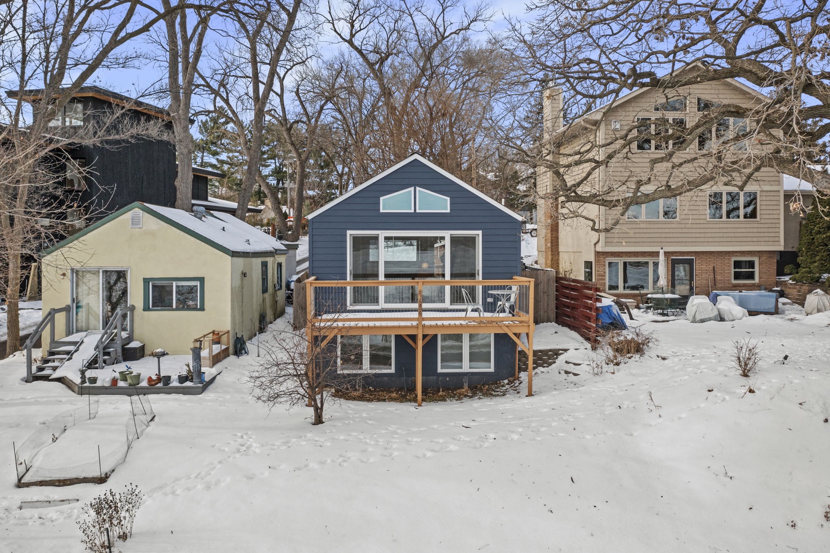 Charming blue cottage nestled in a peaceful winter wonderland, featuring a welcoming deck perfect for cozy seasonal escapes.