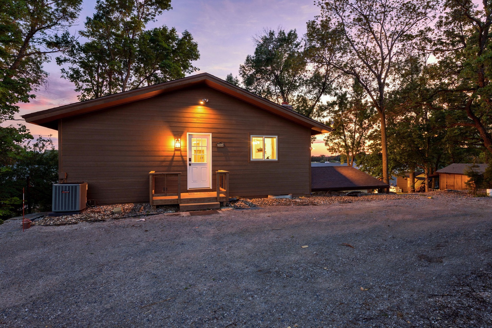 A peaceful woodland cabin nestled among mature trees, offering spacious parking and a welcoming entrance beneath a stunning twilight sky.