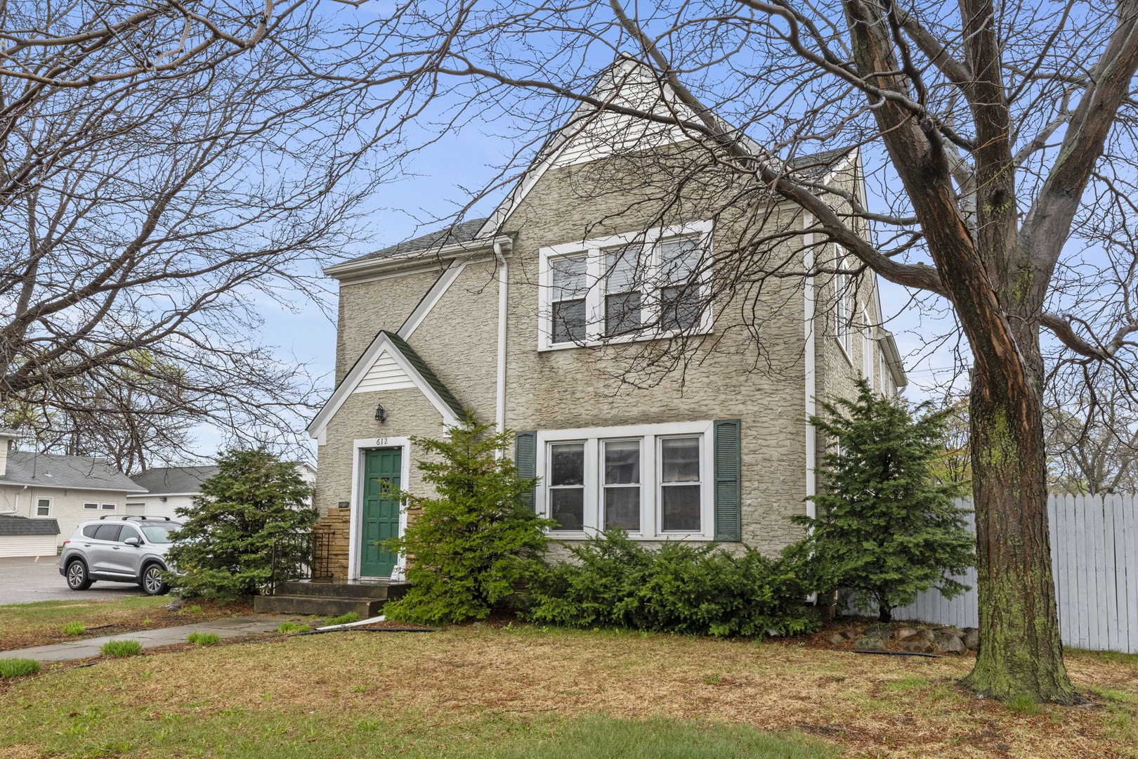 A charming two-story brick home with classic white trim and green shutters, nestled on a quiet residential street surrounded by mature trees and evergreen landscaping.