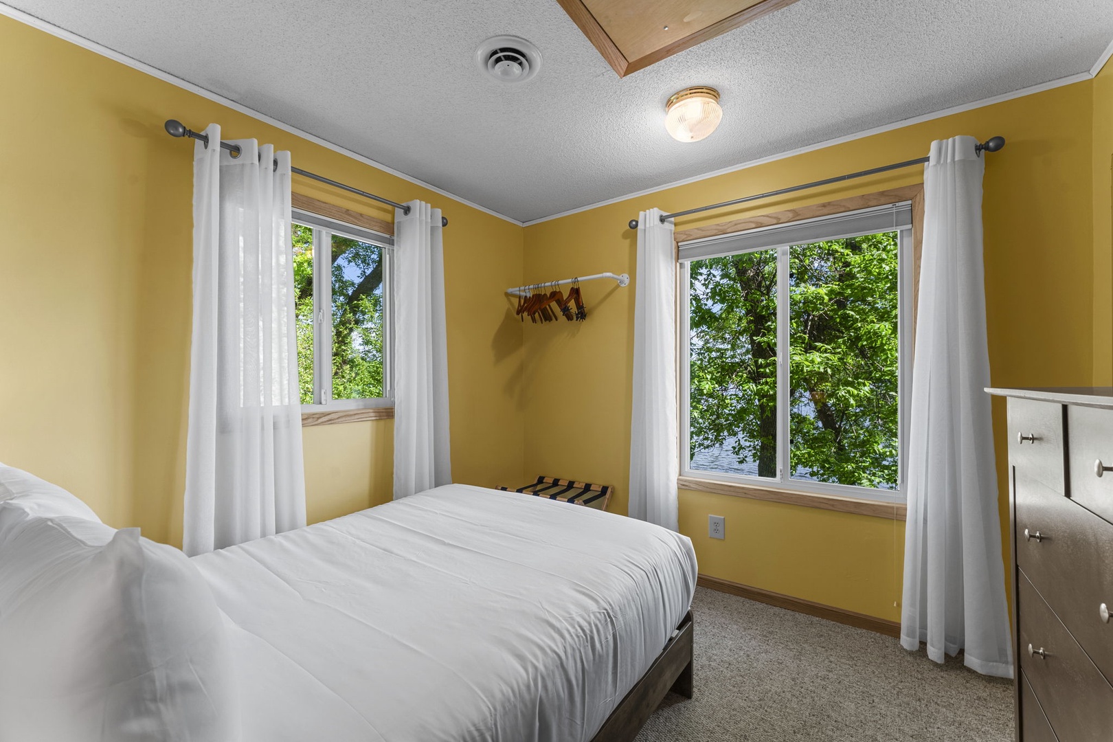 Wake up to sunlight streaming through these bright windows, framing lush green views. Your peaceful retreat features a cozy bed, soft linens, and warm golden tones that invite you to relax and recharge.