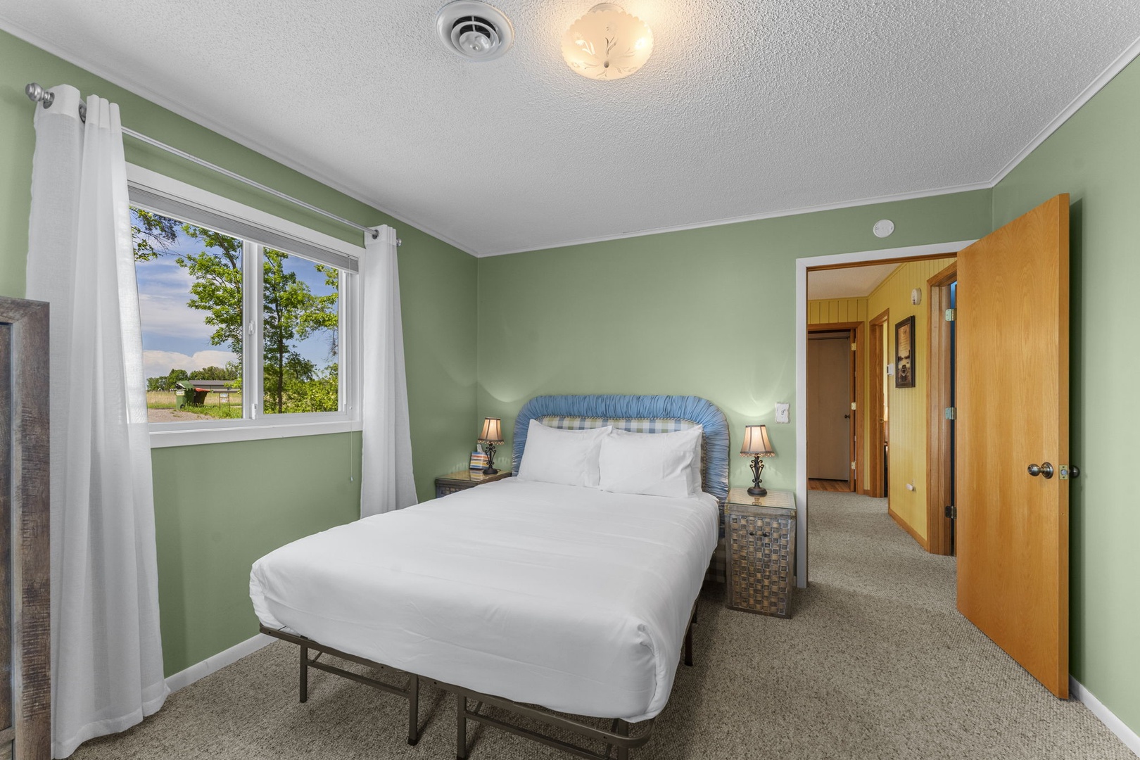 Unwind in your serene bedroom with soothing green walls, crisp white linens, and charming bedside lamps. Natural light streams through the window, revealing peaceful countryside views to start your mornings.