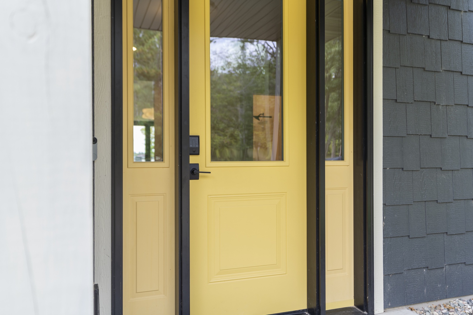 Cheerful yellow front door with modern black trim and sidelights welcomes guests to the property, framed by contemporary gray shingle siding.