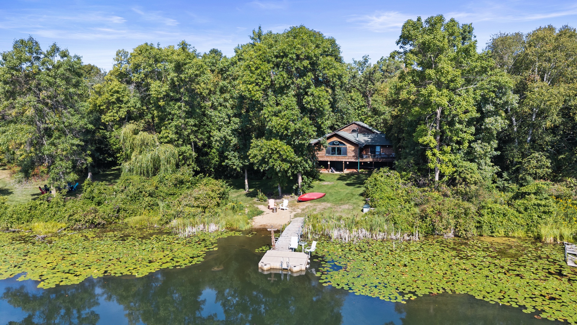A charming lakefront retreat nestled among mature trees, featuring a private dock extending into serene waters dotted with lily pads. The peaceful natural setting offers a perfect escape for those seeking tranquility and outdoor beauty.