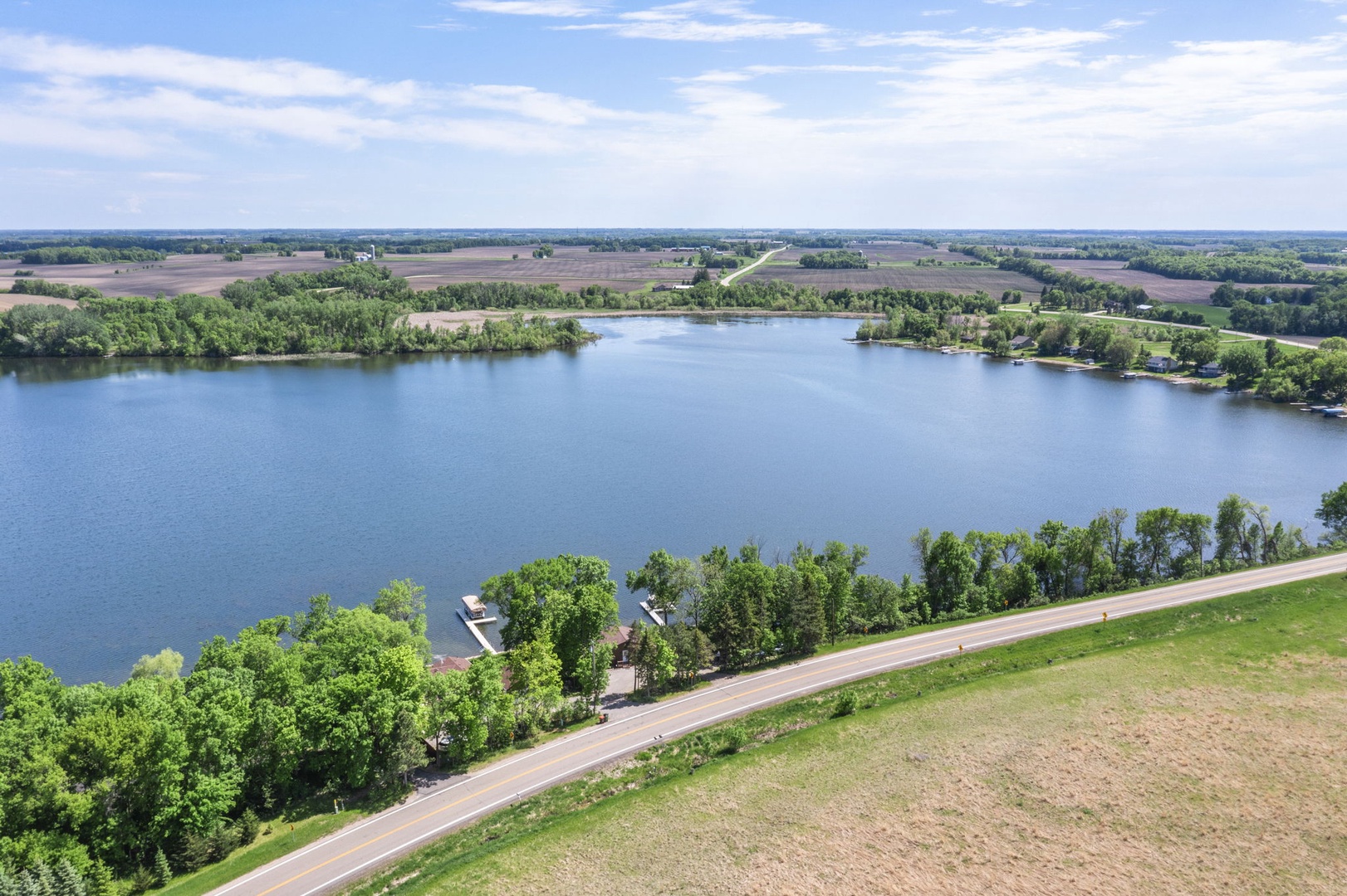 An aerial perspective showcases the property's lakefront setting, with a tranquil lake bordered by lush greenery, rolling farmland, and a quiet rural road winding through the scenic countryside.