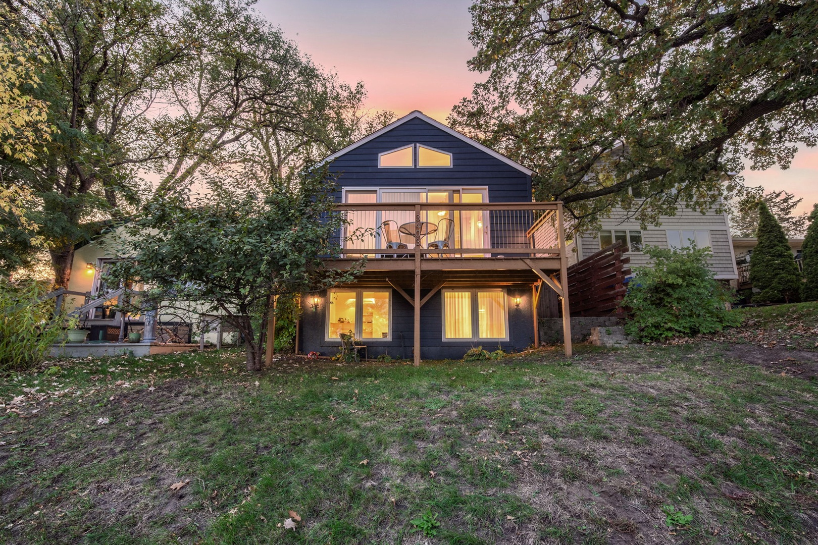 The property sits peacefully among mature trees, with warm lights glowing from within as sunset paints the sky in soft hues, welcoming guests to their waterfront retreat.
