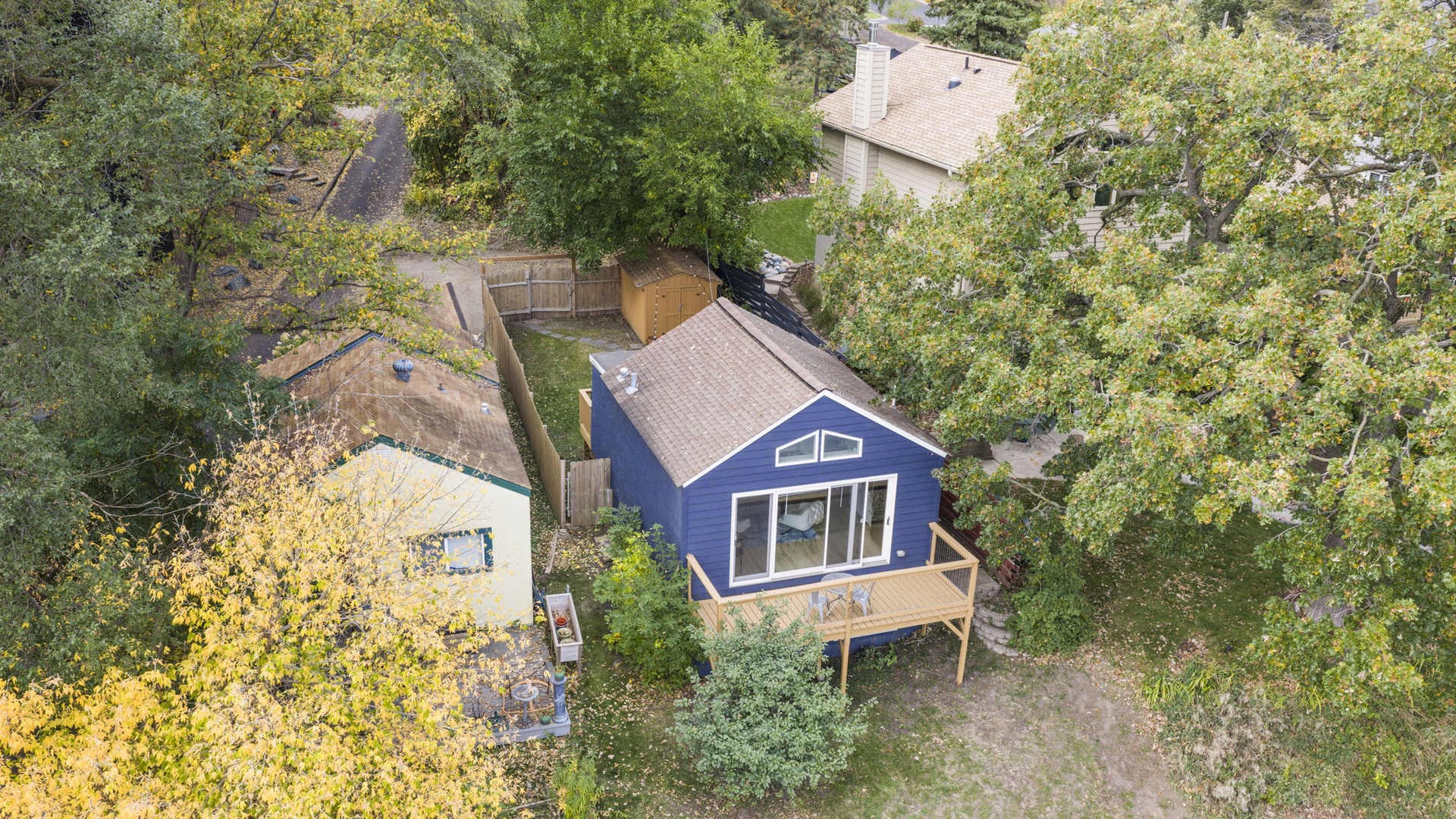 Aerial view of a charming blue cottage nestled among mature trees in a quiet residential area, featuring a spacious deck and private fenced yard.