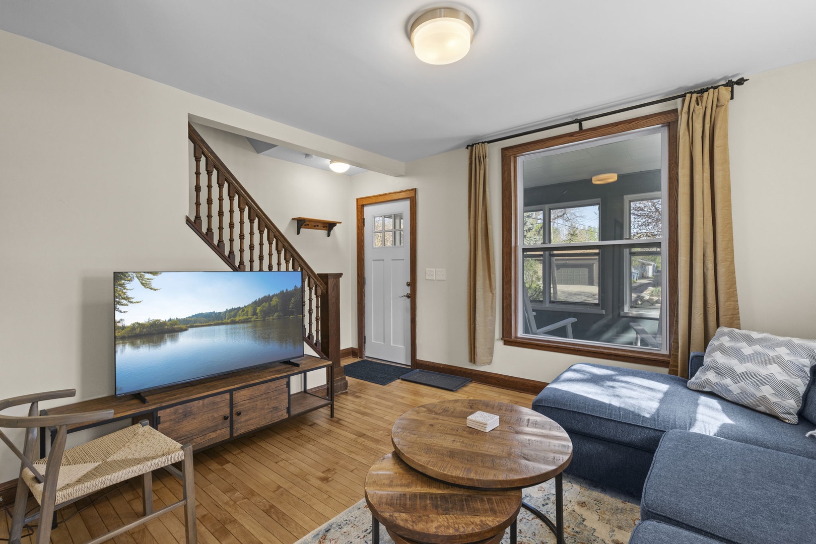 Spacious living room with large smart TV, staircase, and front entry door — hardwood floors and big windows keep the space bright and inviting all day.