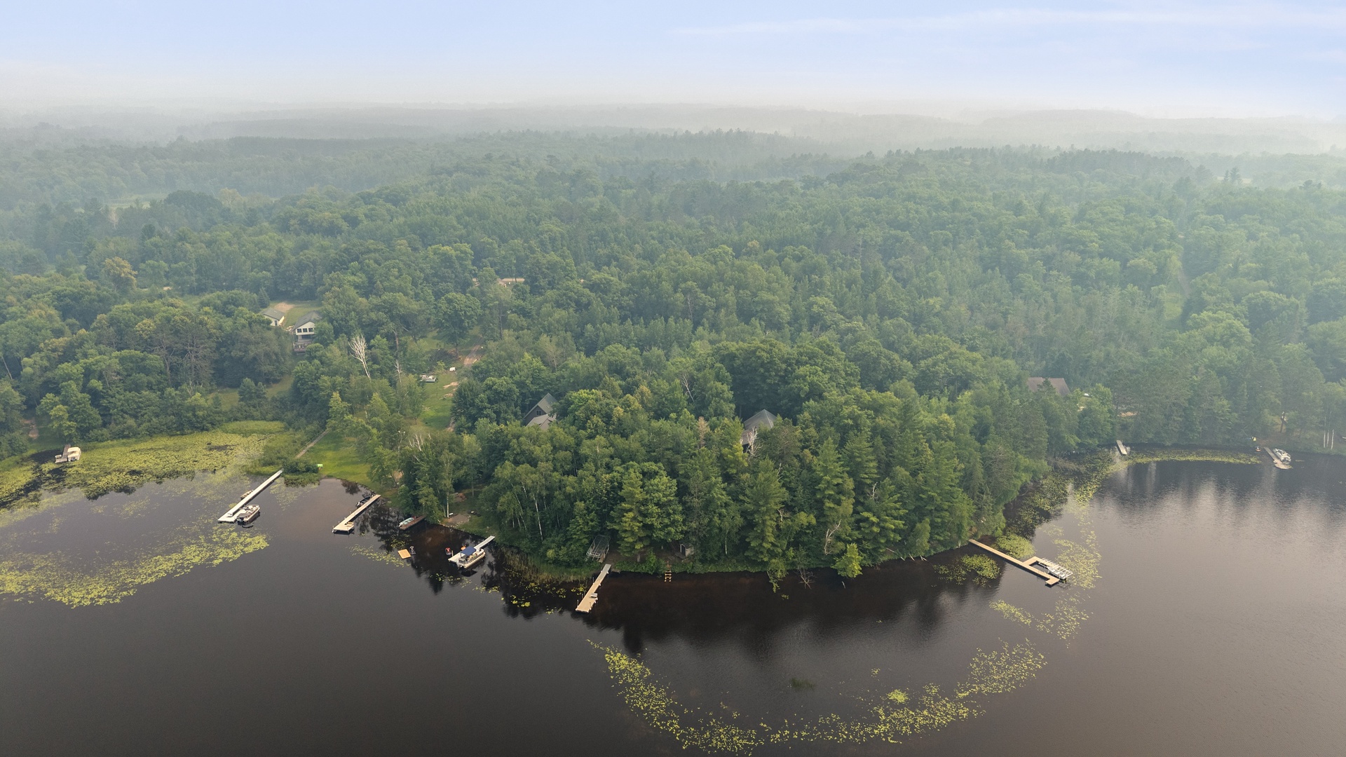 Aerial view of a secluded lakefront property nestled in pristine forest, with private docks extending into calm waters surrounded by lush natural wilderness.
