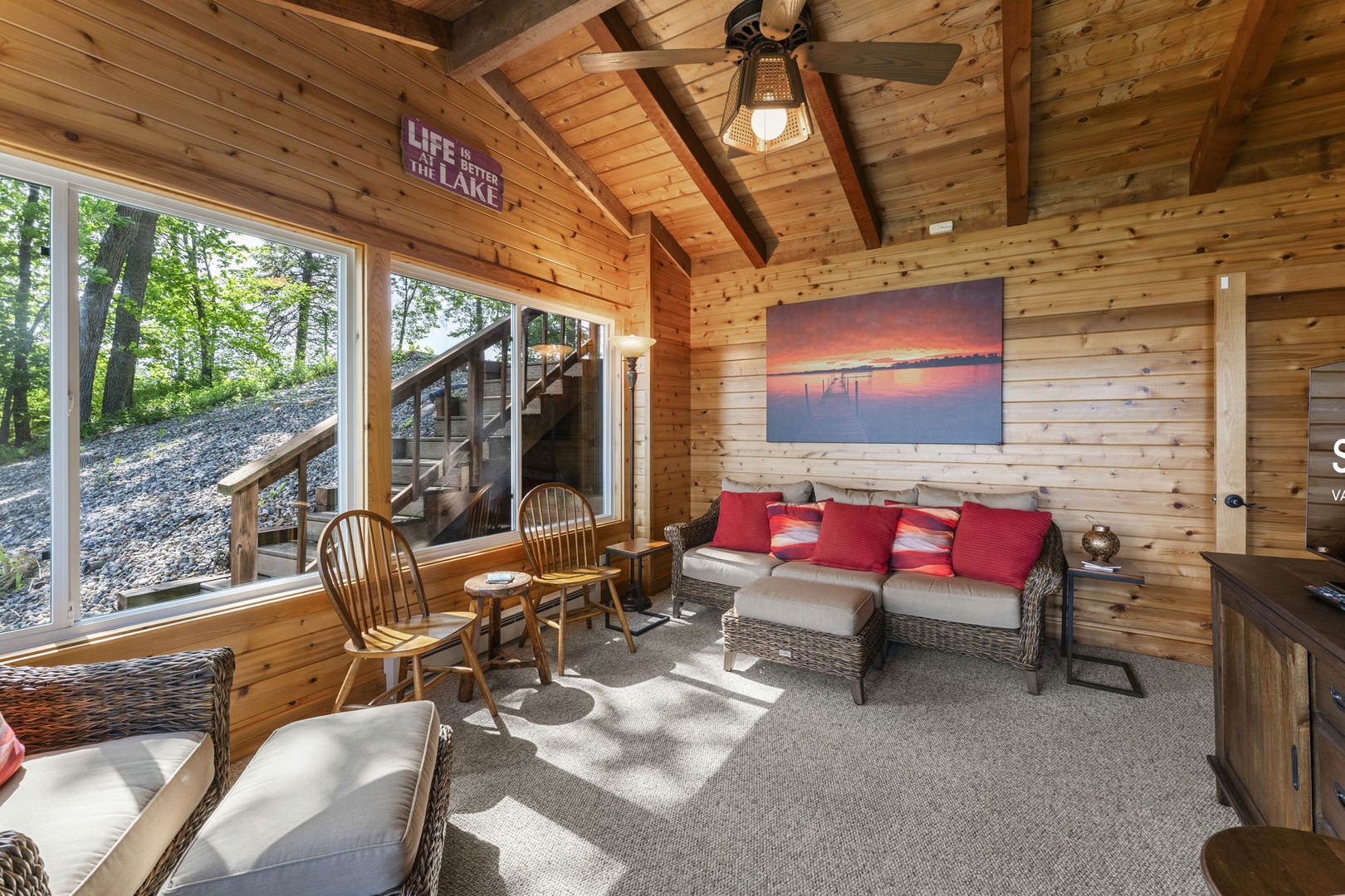 Step into this warm, light-filled living room where rustic wood meets lakeside comfort. Settle into the wicker seating, gaze at the peaceful sunset art, and enjoy nature's beauty through large windows that frame the wooded landscape.