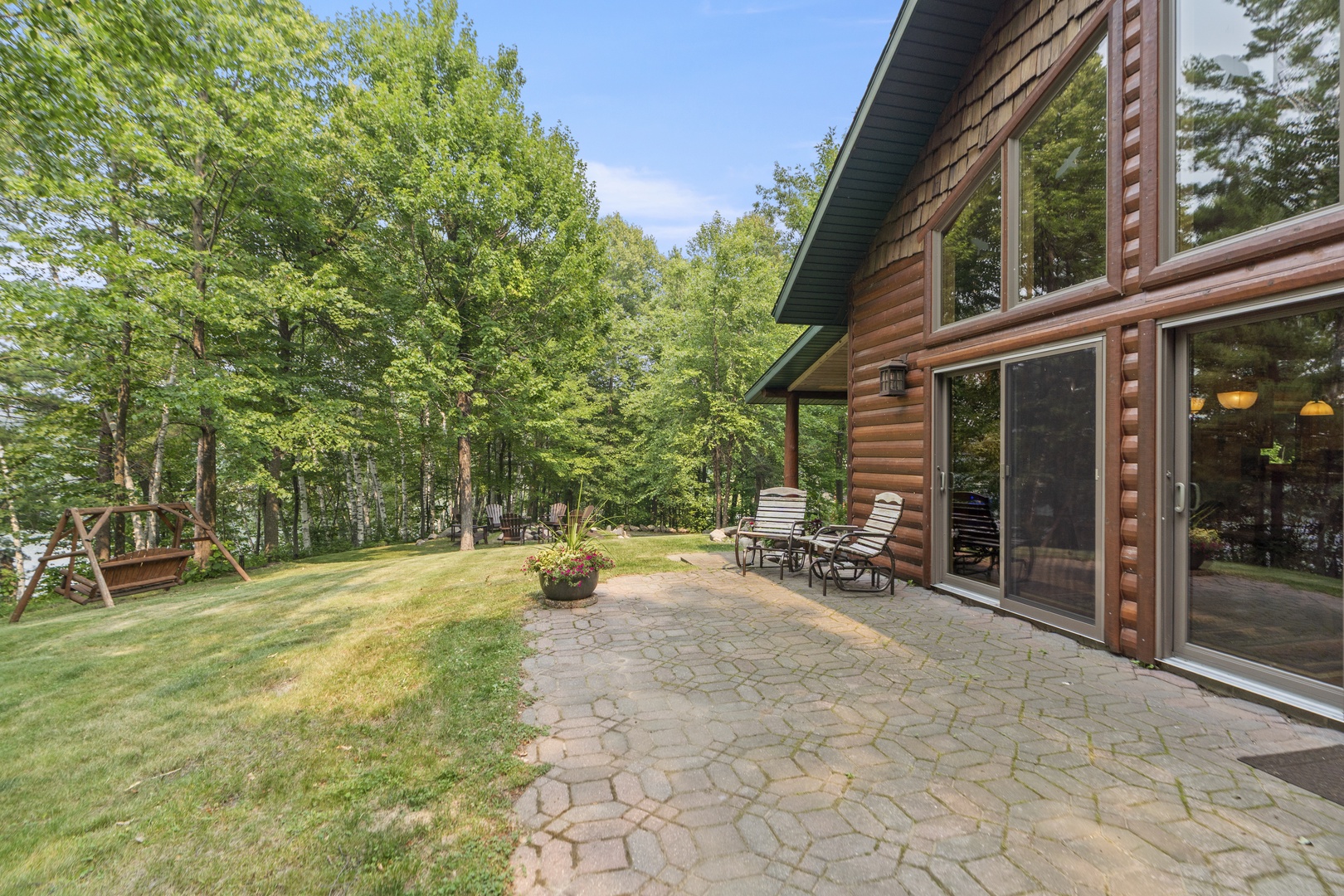 A charming log cabin nestled among tall trees, featuring a spacious stone patio where you can unwind in the fresh air. Lush green lawn and forest views create a peaceful, private retreat.