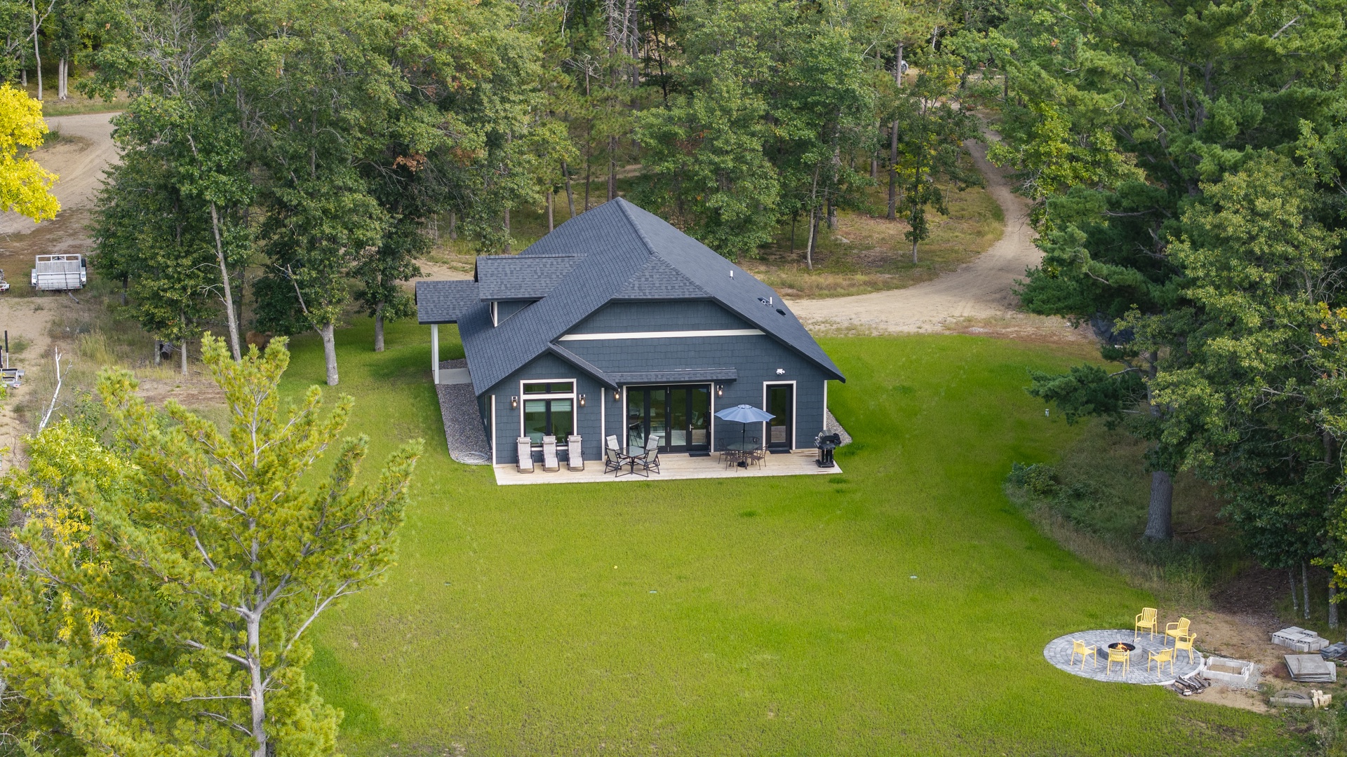 A modern vacation home with covered patio and outdoor seating sits peacefully among towering trees, with a spacious lawn and firepit area perfect for summer evenings.