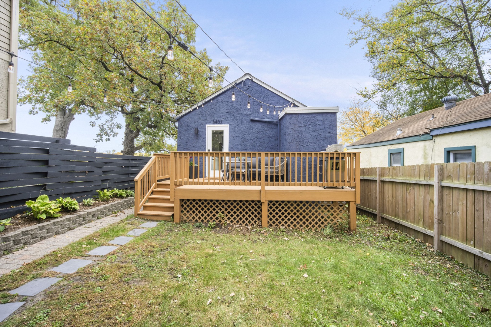 The property features a private backyard with a spacious deck, grassy lawn, and landscaped garden areas surrounded by mature trees. The charming blue building serves as part of the rental's outdoor setting.