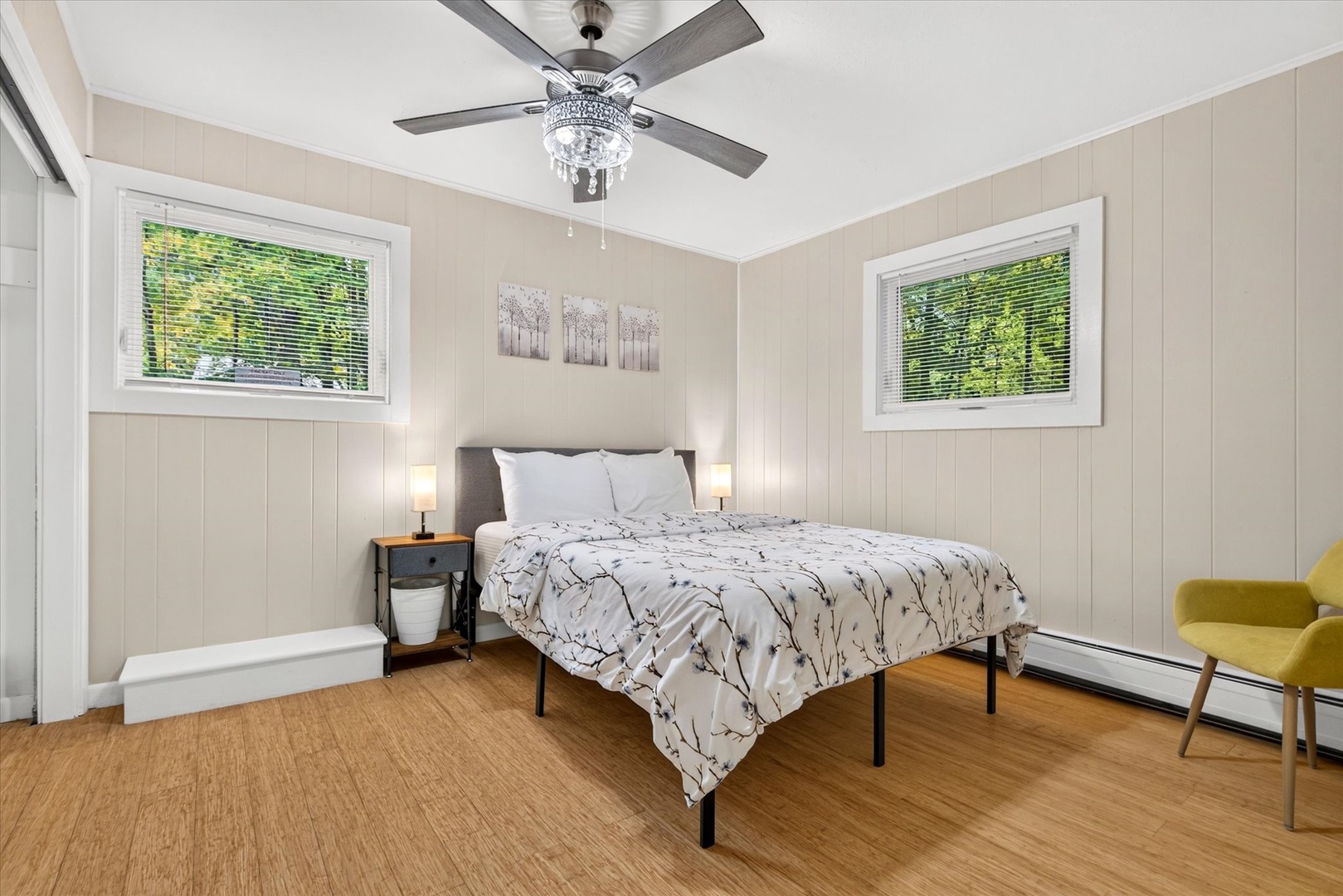 Drift off in your peaceful bedroom, where soft natural light filters through two large windows and a sparkling chandelier ceiling fan adds a touch of elegance overhead.