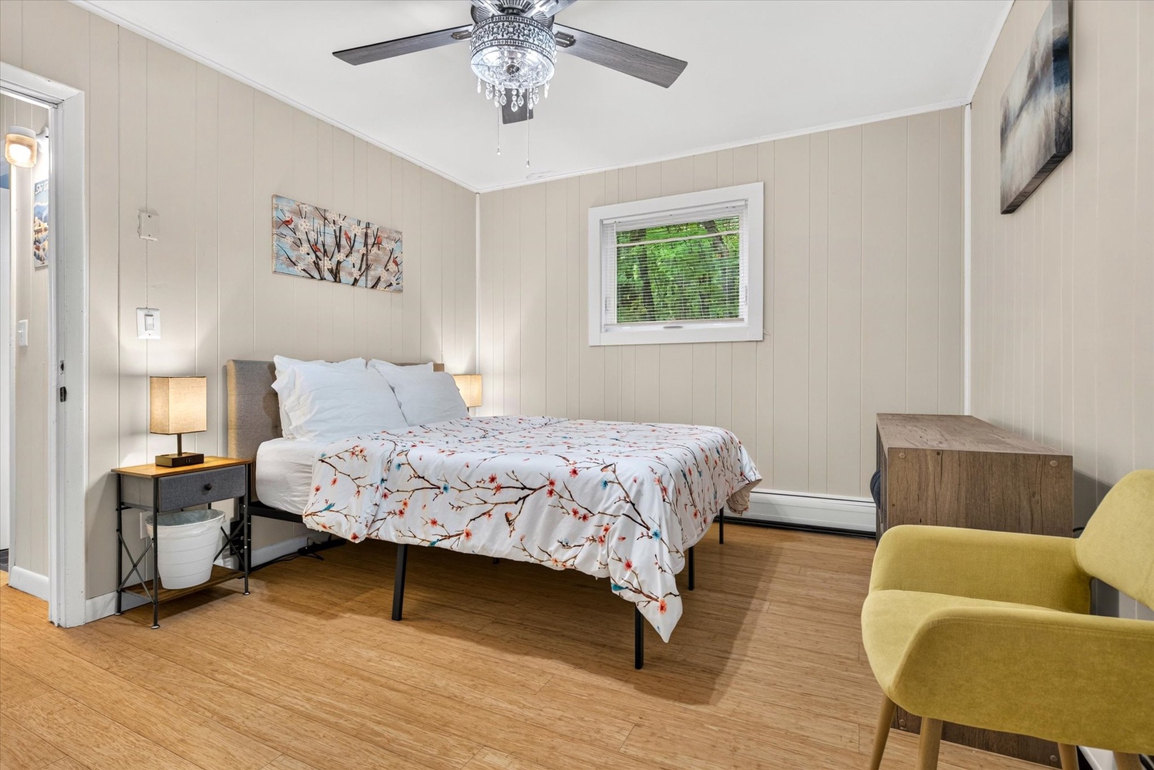 Your peaceful retreat features crisp linens, soft lighting, and a gentle ceiling fan to keep you comfortable. A cozy chair invites you to unwind with a book, while natural light filters through the window overlooking lush greenery.