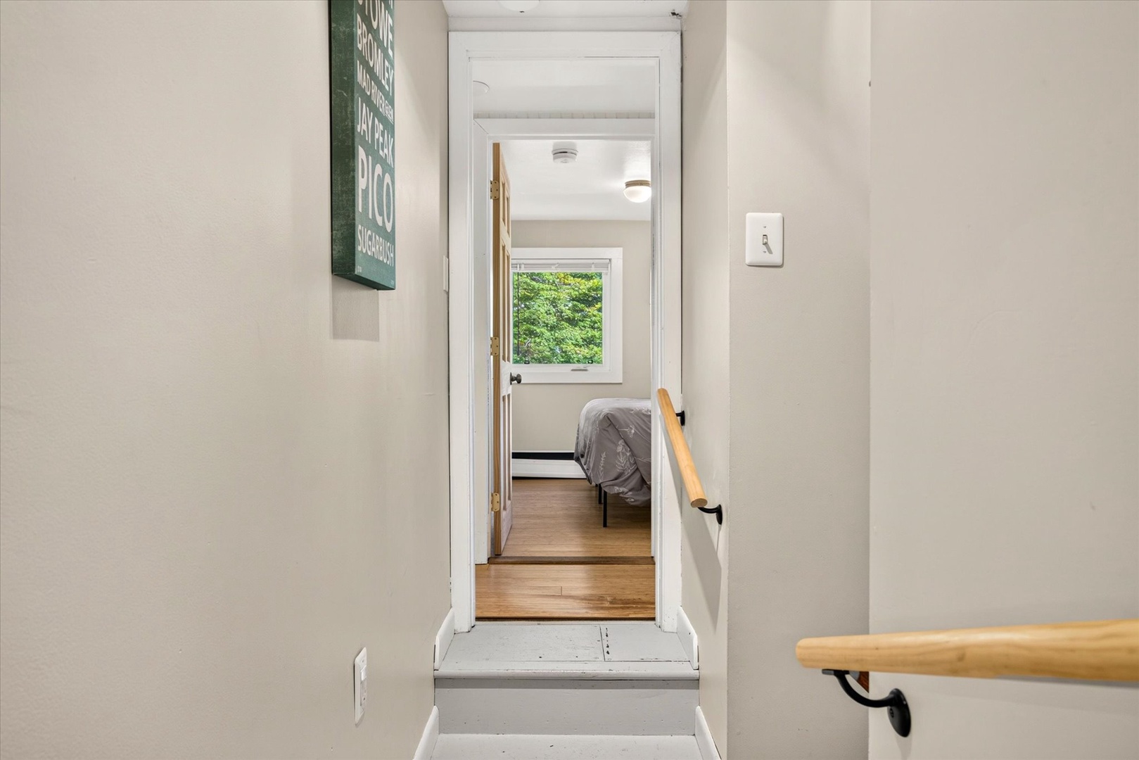 A cozy bedroom awaits at the top of the stairs, with natural light streaming through the window and lush greenery framing your peaceful retreat.
