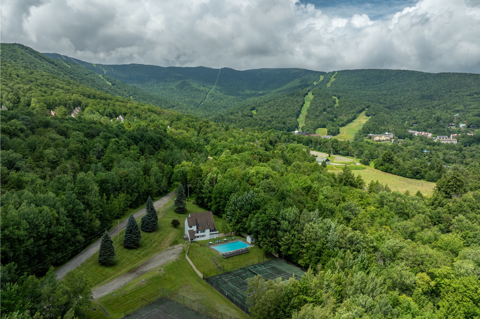 An aerial view captures the property nestled in lush mountain scenery, with a pool, tennis court, and access to nearby slopes.