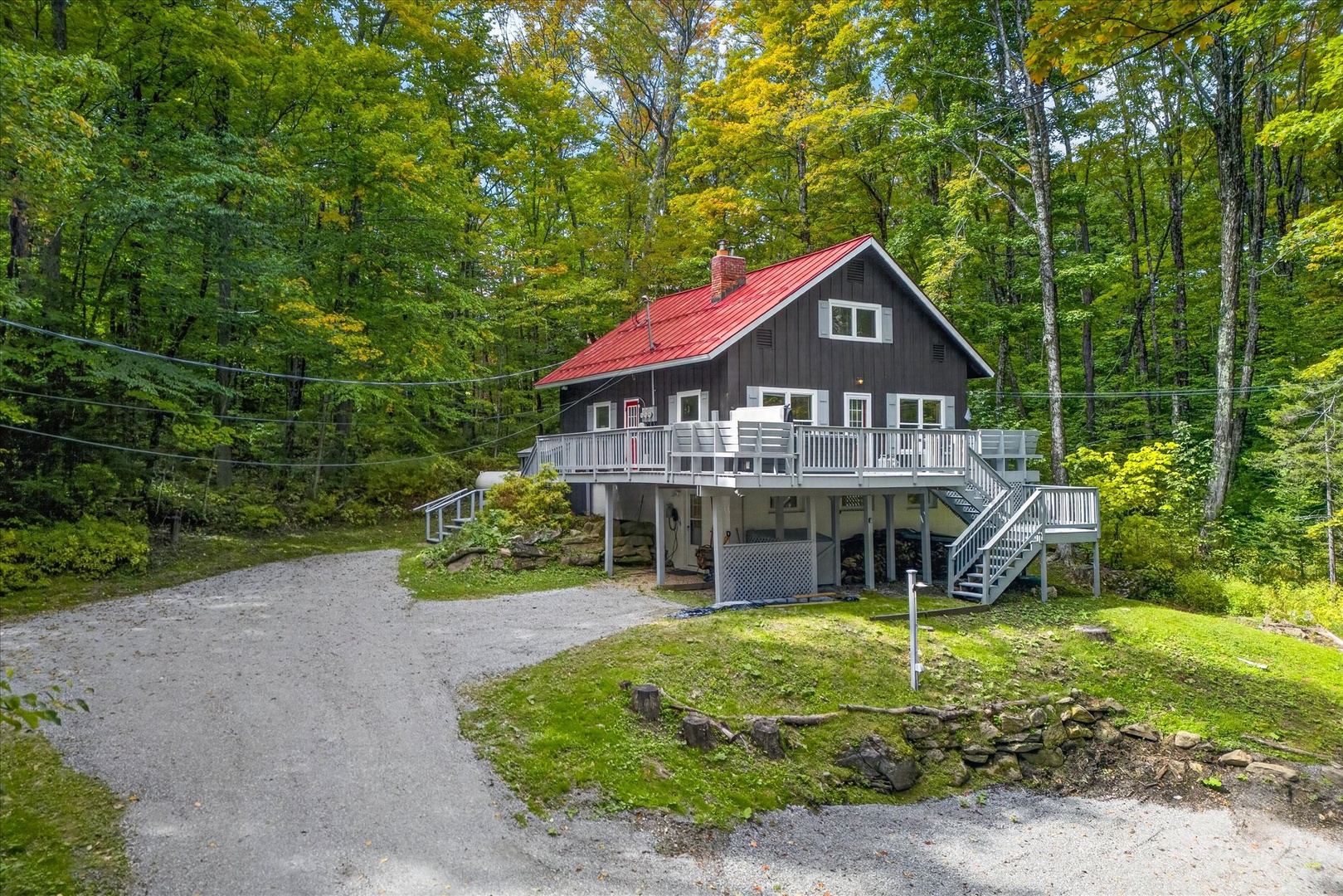 This charming forest retreat features a distinctive red roof and wraparound deck nestled among vibrant autumn foliage, with convenient parking and easy access to the main entrance.
