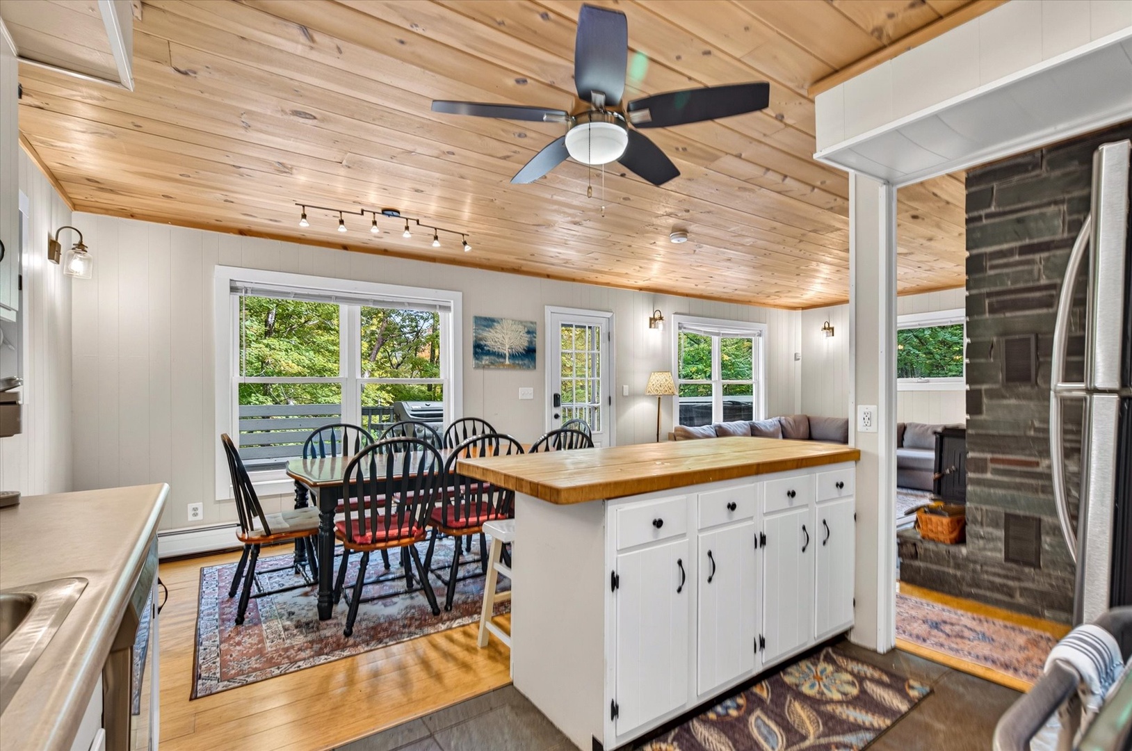 Gather around the rustic dining table under warm wood-paneled ceilings, where natural light spills through large windows and the kitchen island invites you to share meals together in this welcoming open-plan space.