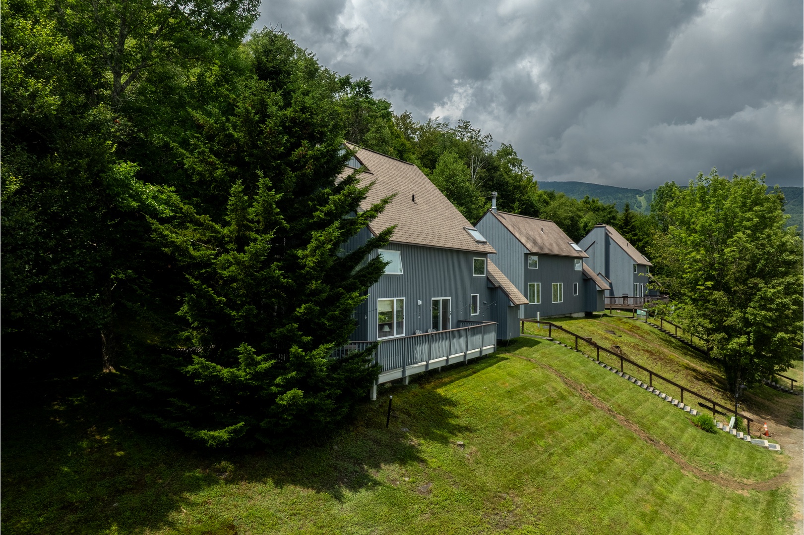 Modern mountain retreat nestled in lush greenery with dramatic storm clouds rolling over distant peaks, offering a tranquil escape surrounded by nature's beauty.