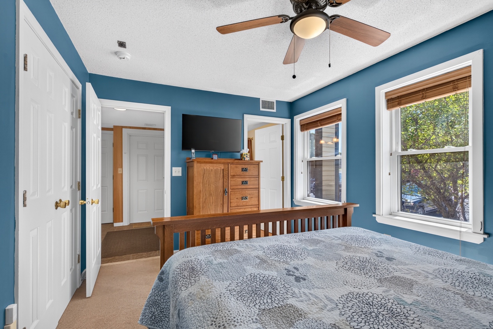 Sink into your peaceful retreat with soothing blue walls, a plush bed, and natural light streaming through large windows that frame the leafy views outside.