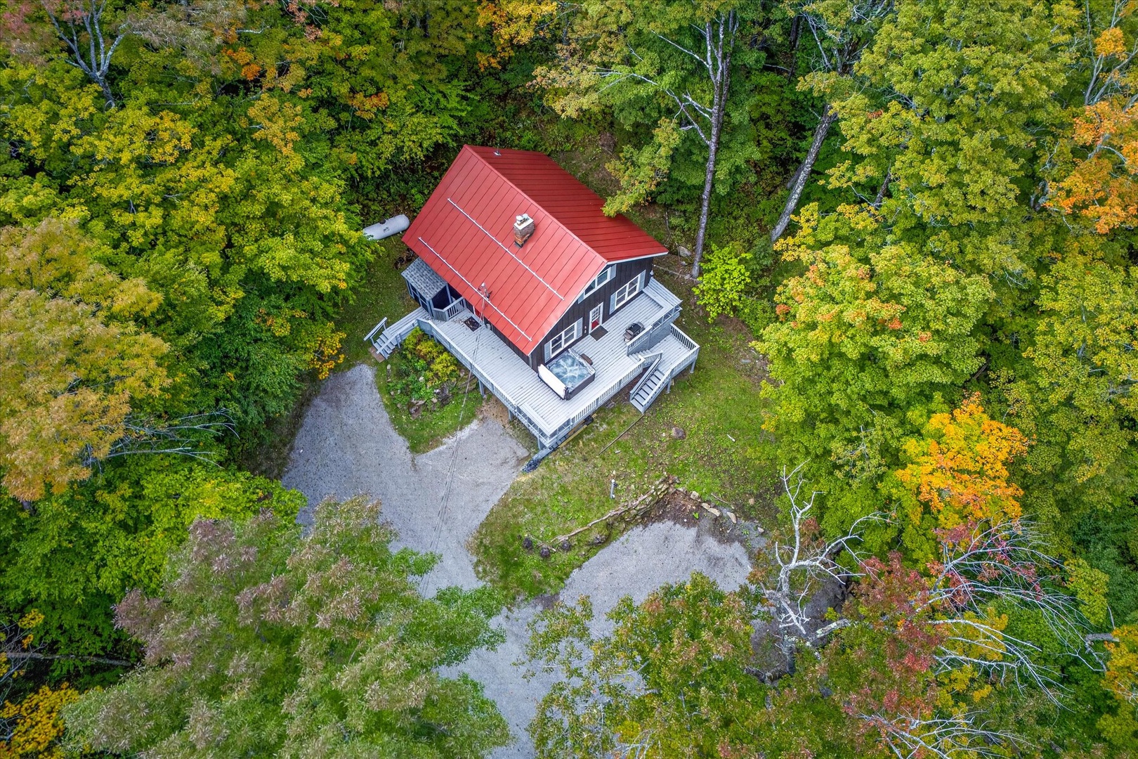 An aerial view captures a charming property with a distinctive red roof nestled in a forest setting during autumn, with vibrant fall foliage painting the surrounding landscape in brilliant greens, golds, and oranges.
