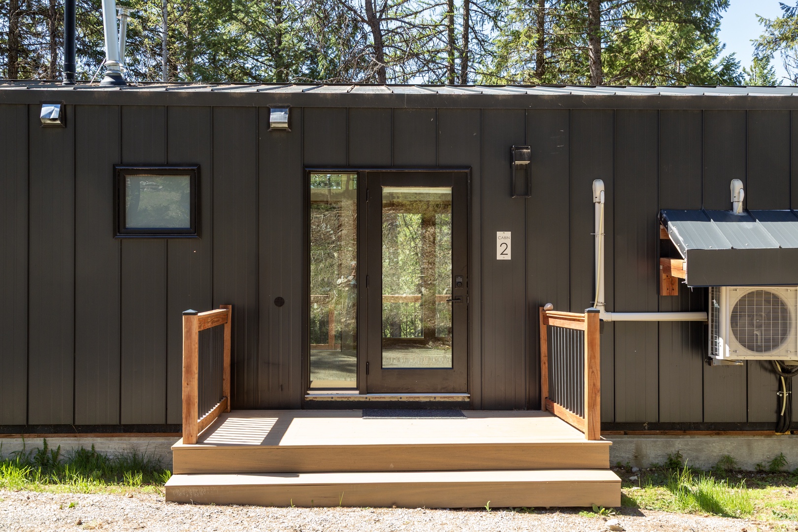 Modern cabin entrance featuring dark siding, glass door entry, and woodland setting—your peaceful retreat awaits with climate-controlled comfort and natural surroundings.