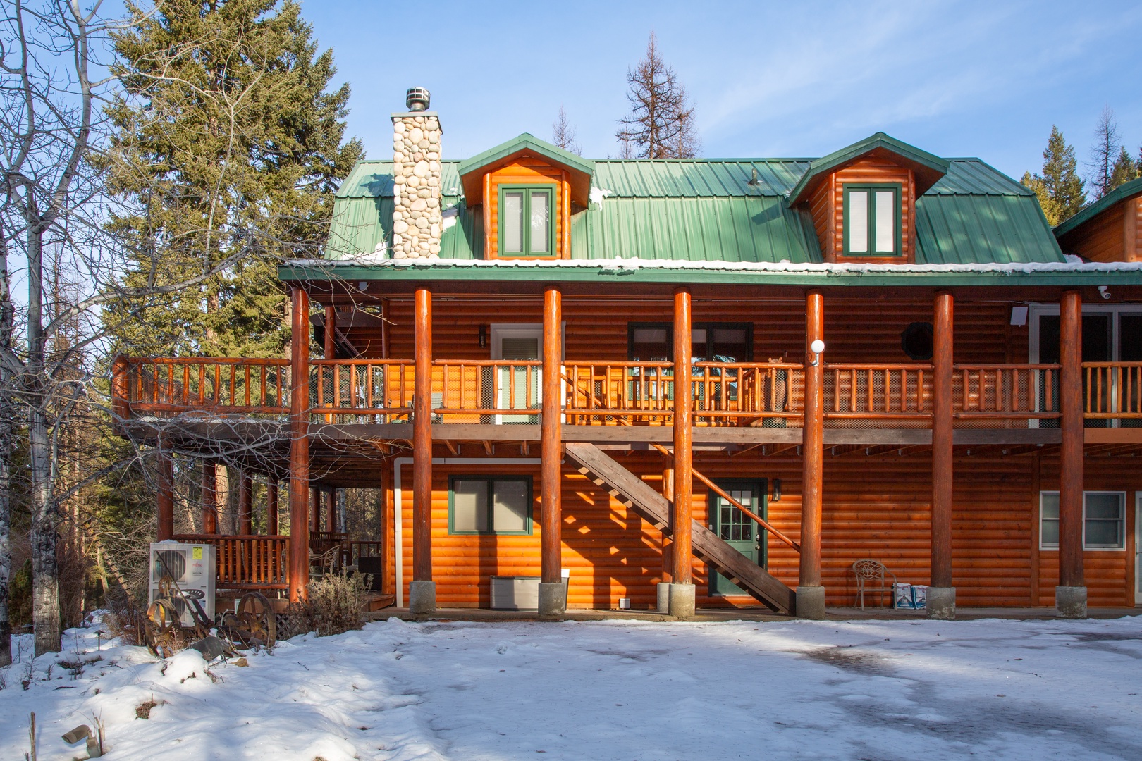 Charming log cabin retreat with green metal roof and spacious wraparound deck, nestled among evergreens with light snow dusting the ground.