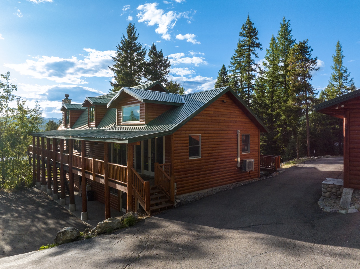A spacious log cabin with a green metal roof sits nestled among towering pines. The multi-level layout, expansive deck, and mountain setting create the perfect retreat for relaxation.