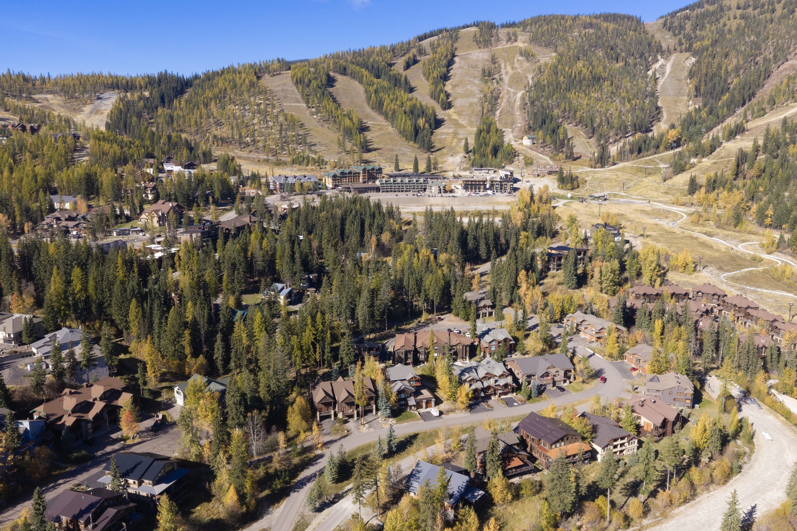 An aerial view captures a mountain resort community nestled among evergreens and autumn foliage, with ski runs visible on the surrounding peaks.