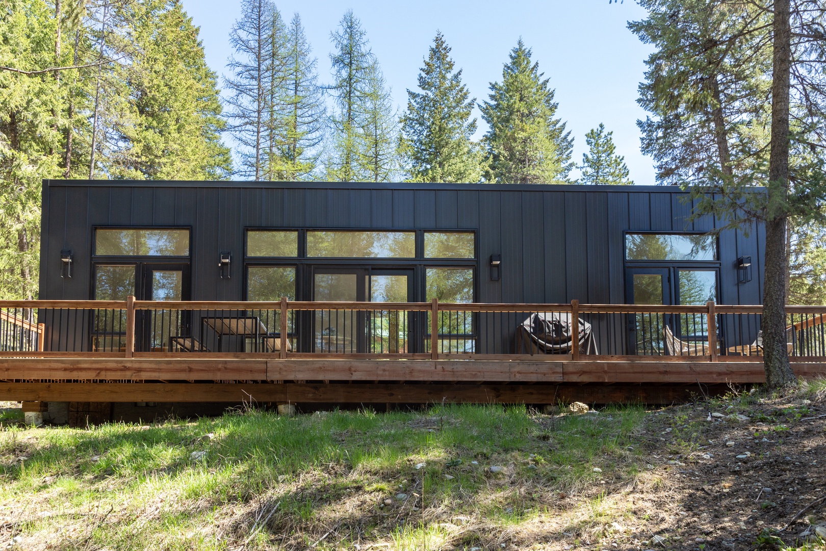 Modern forest retreat featuring sleek black vertical siding and expansive windows framed by towering evergreens. The wrap-around wooden deck offers a peaceful outdoor space for relaxation.