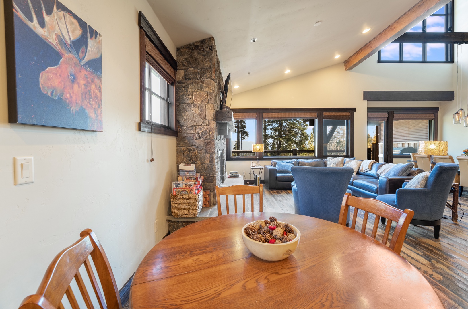 Spacious living area with exposed timber beams, stone fireplace, and floor-to-ceiling windows framing sweeping mountain views. Plush blue sofas invite relaxation in this warm retreat bathed in natural light.