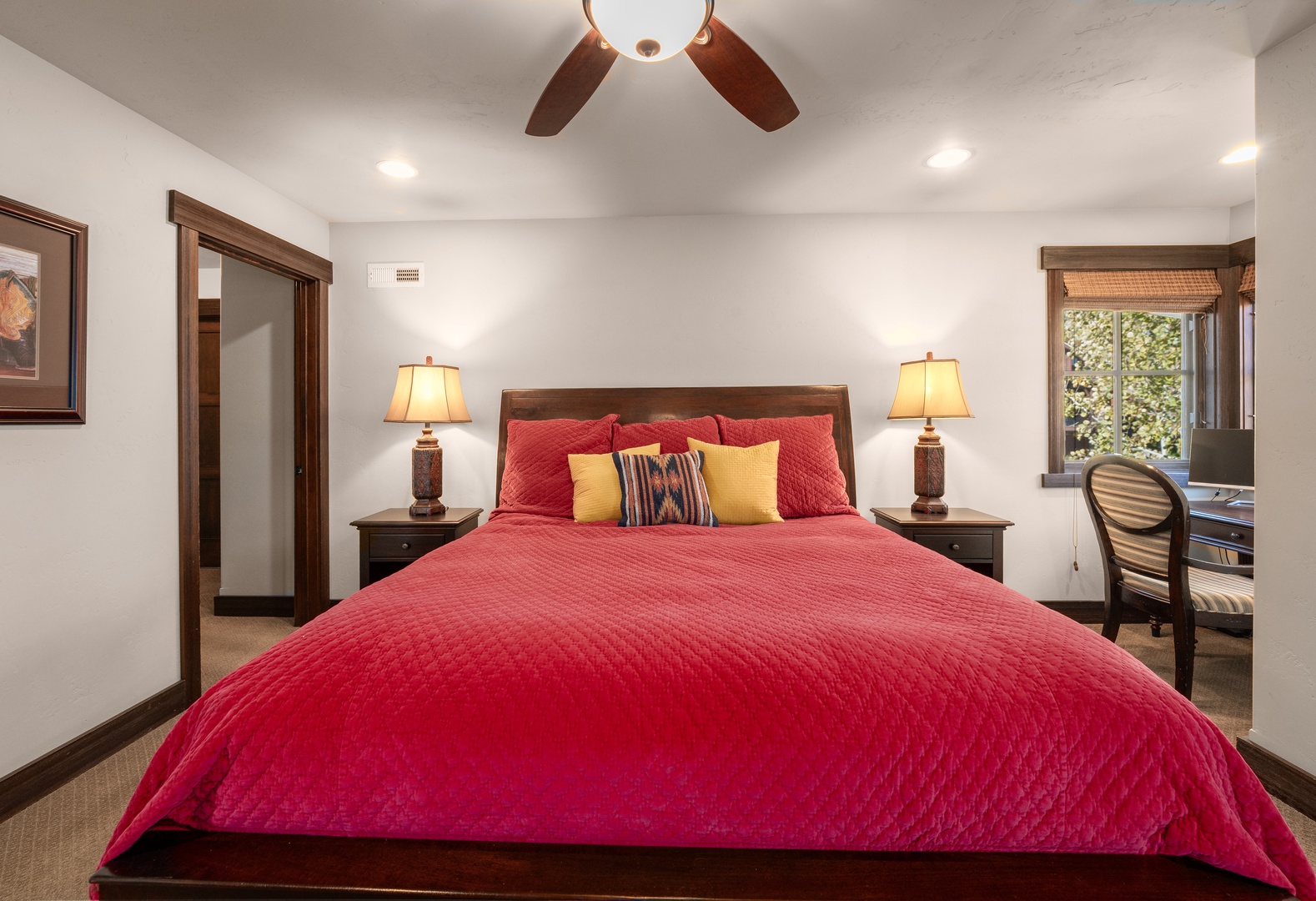 Sink into the plush bed dressed in inviting crimson tones, while warm lamps cast a soft glow across your peaceful retreat. A ceiling fan keeps the air just right, and a quiet desk awaits your morning coffee or evening reflection.