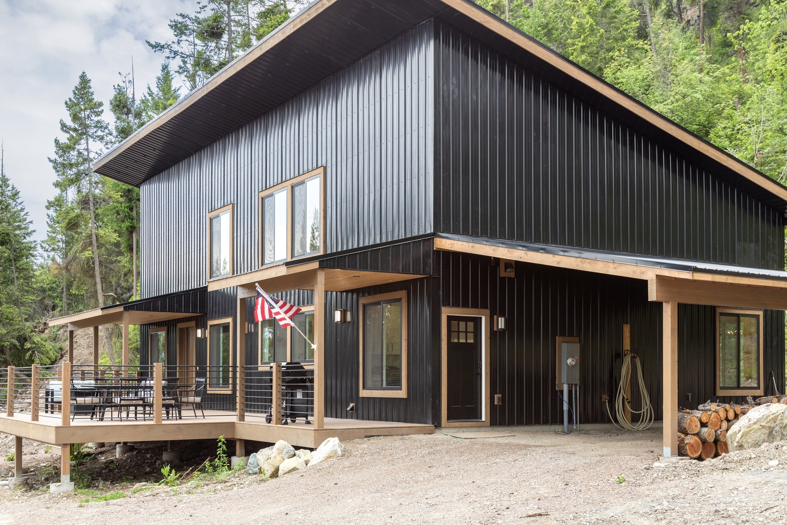 Modern two-story vacation rental features striking black corrugated siding, warm wood accents, and a spacious deck nestled among towering evergreens. Gravel driveway provides easy access to this secluded forest retreat.
