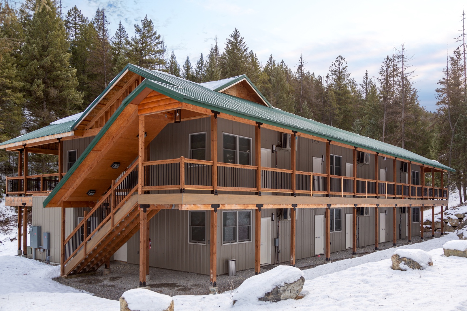 Nestled among towering pines in a peaceful winter setting, this rustic mountain lodge features a green metal roof and wraparound balconies surrounded by scenic forestland.