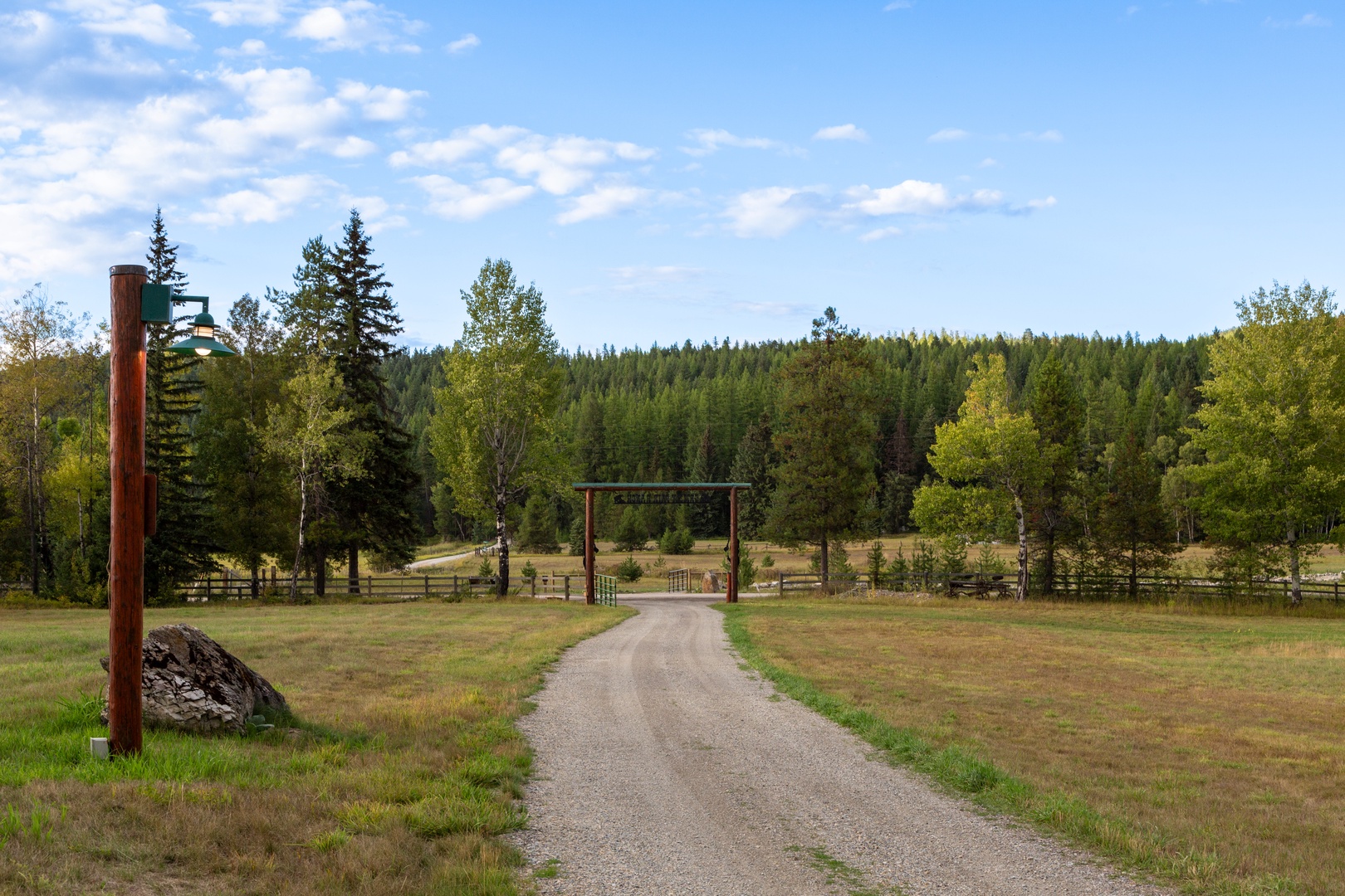 A peaceful gravel driveway leads through rolling countryside to the property entrance, surrounded by dense forest and open meadows.