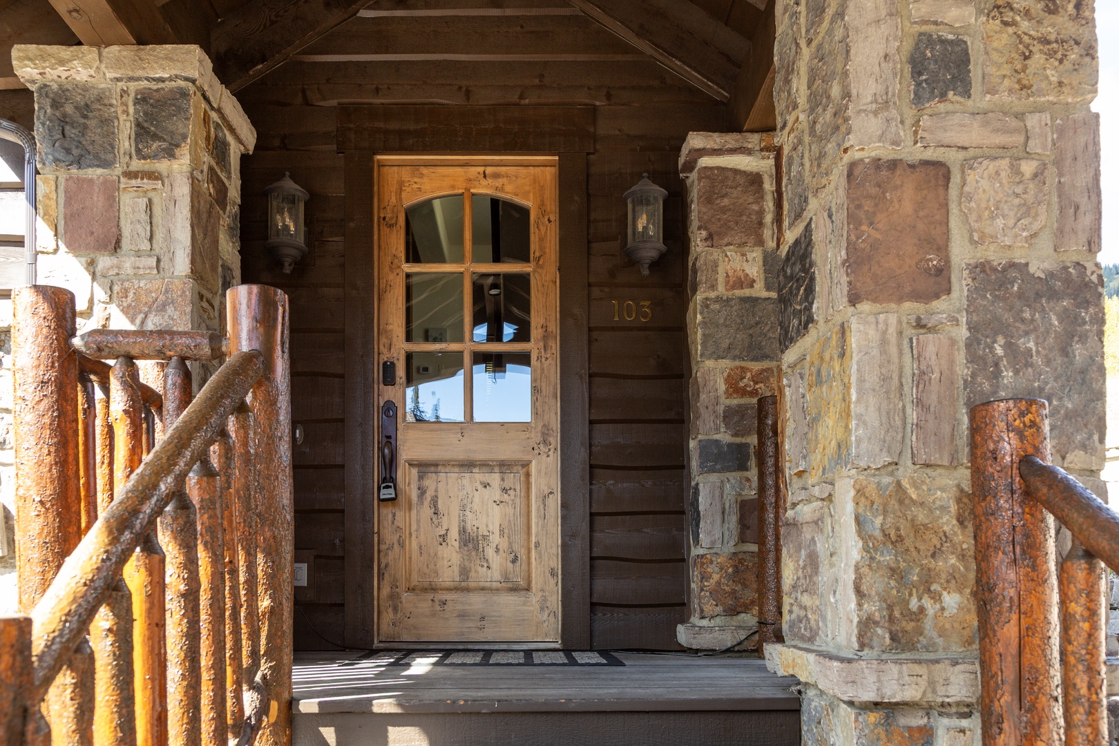 Rustic mountain lodge entrance featuring reclaimed wood, natural stone pillars, and handcrafted log railings marking your arrival to an authentic alpine retreat.