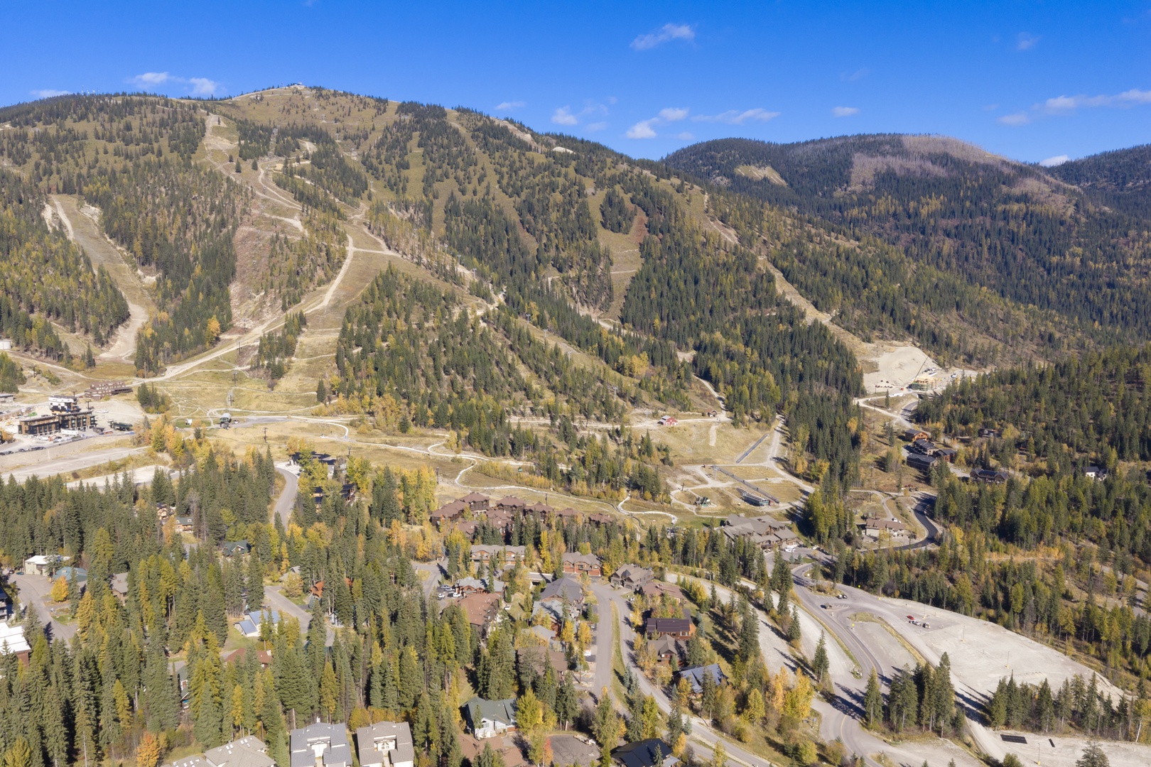 This aerial view showcases a picturesque mountain resort area with forested slopes, ski runs, and winding roads through evergreen and autumn-tinged trees.