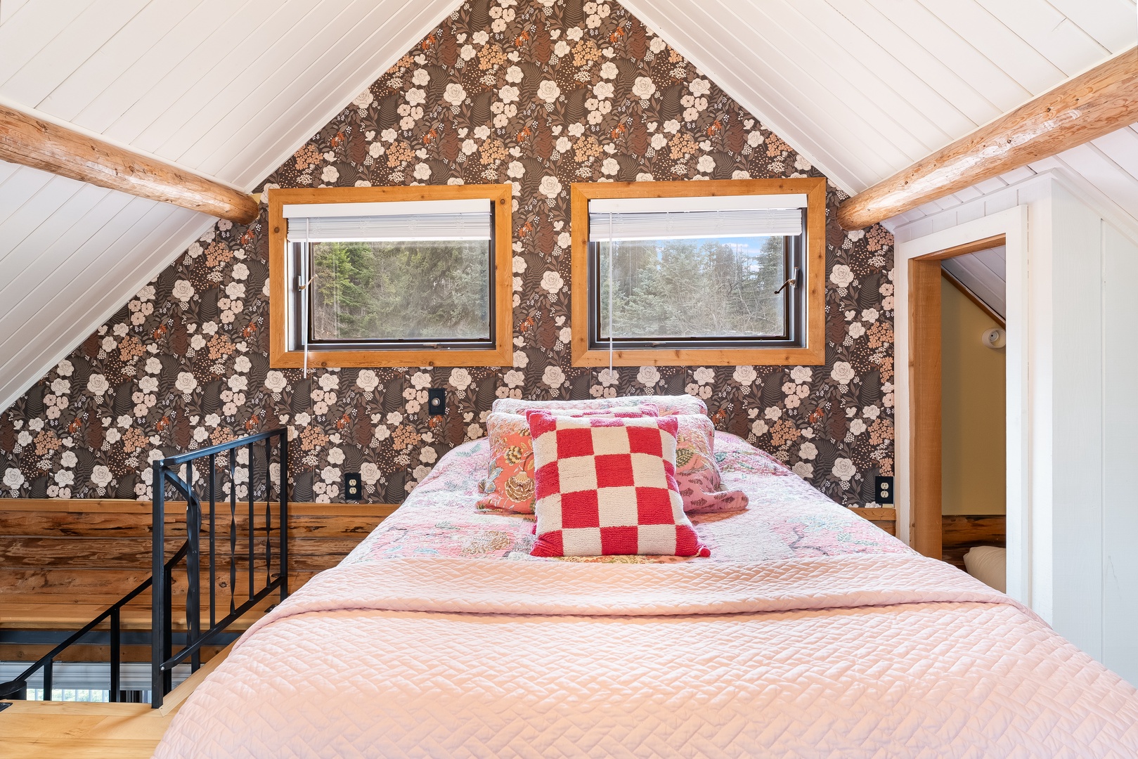 Your cozy loft bedroom features charming vintage wallpaper, warm wood beams, and peaceful forest views through twin windows.
