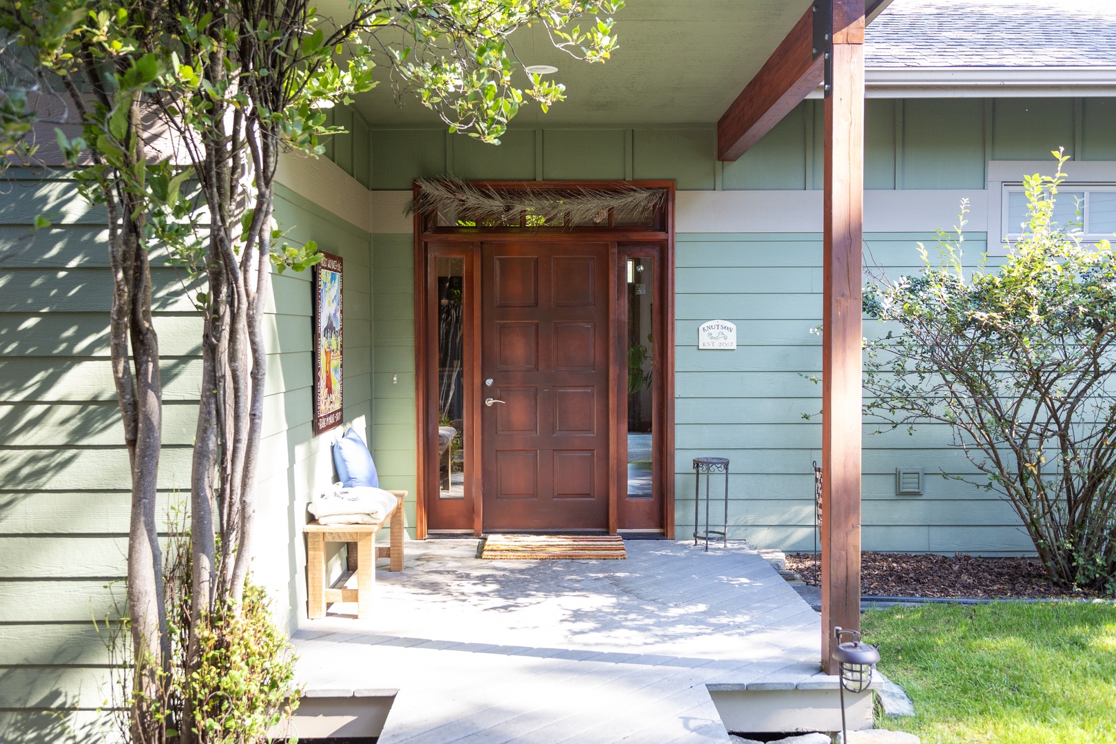 This charming sage-green façade features a welcoming entryway framed by natural wood accents and lush greenery, setting a serene tone for your arrival.