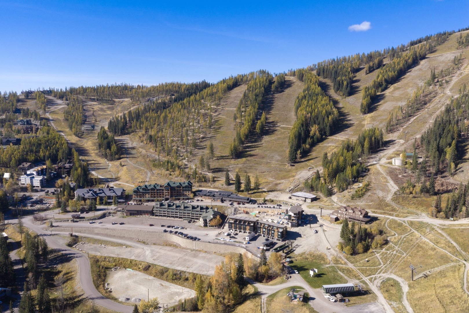 An aerial view captures the property nestled at the base of a scenic mountain ski area, surrounded by evergreen forests and open terrain under a clear blue sky.