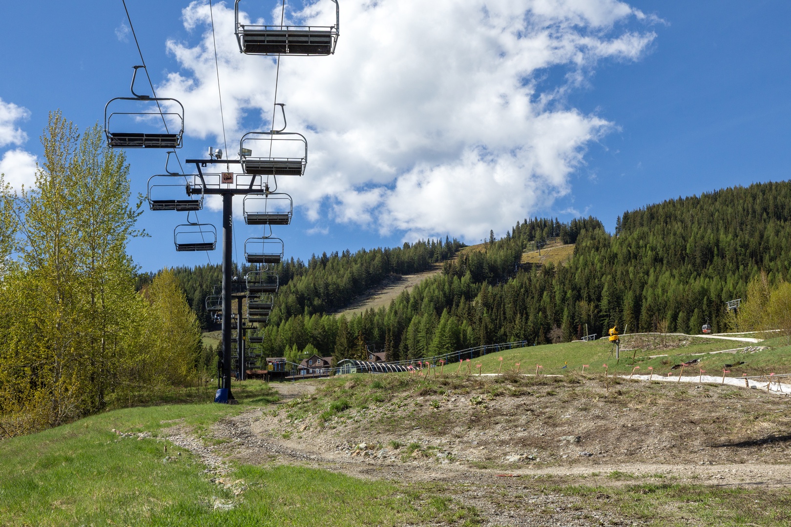 A ski resort area featuring chairlifts ascending pine-covered slopes, with off-season trails and facilities visible beneath dramatic blue skies and billowing white clouds.