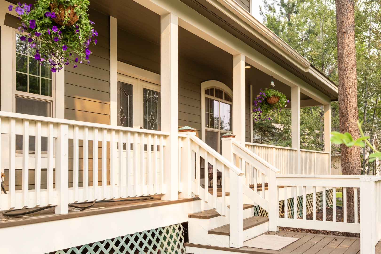 A welcoming covered front porch surrounded by lush greenery and tall pines, with vibrant hanging flower baskets framing the entry steps.