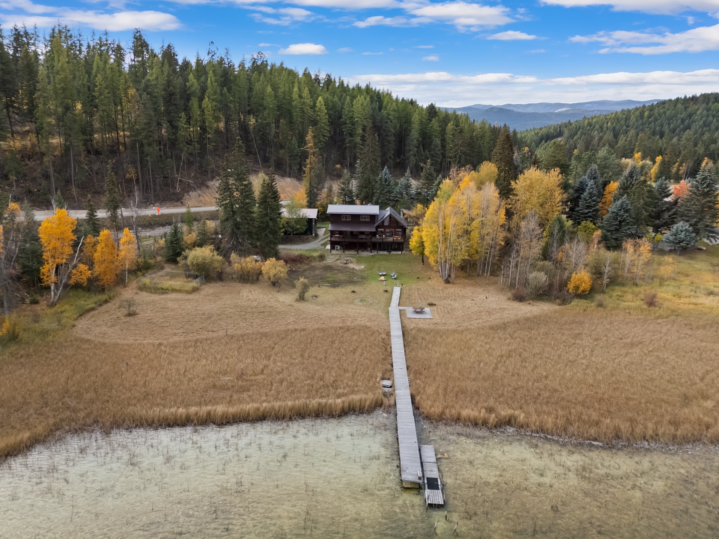 Aerial view of the property nestled among evergreen forests and autumn foliage, featuring a private dock extending into tranquil waters.