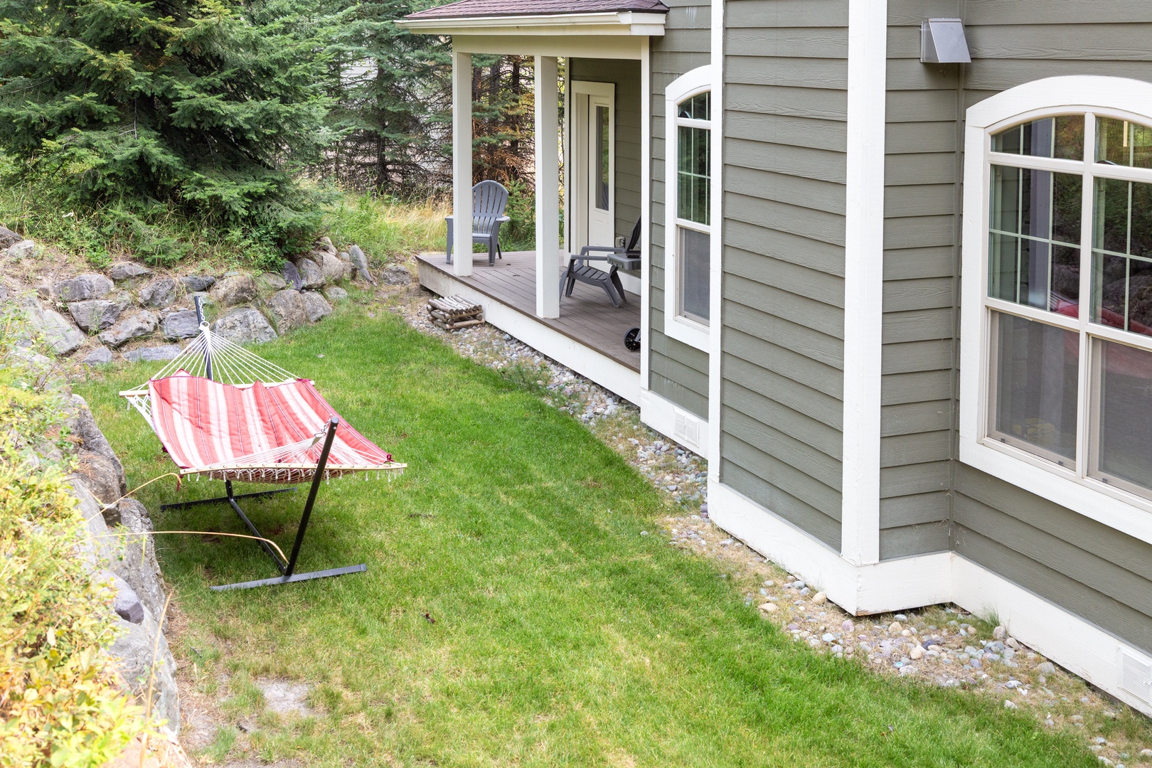 Your private backyard oasis features a striped hammock on lush lawn, perfect for lazy afternoons surrounded by natural rock outcroppings and a covered porch with Adirondack seating.